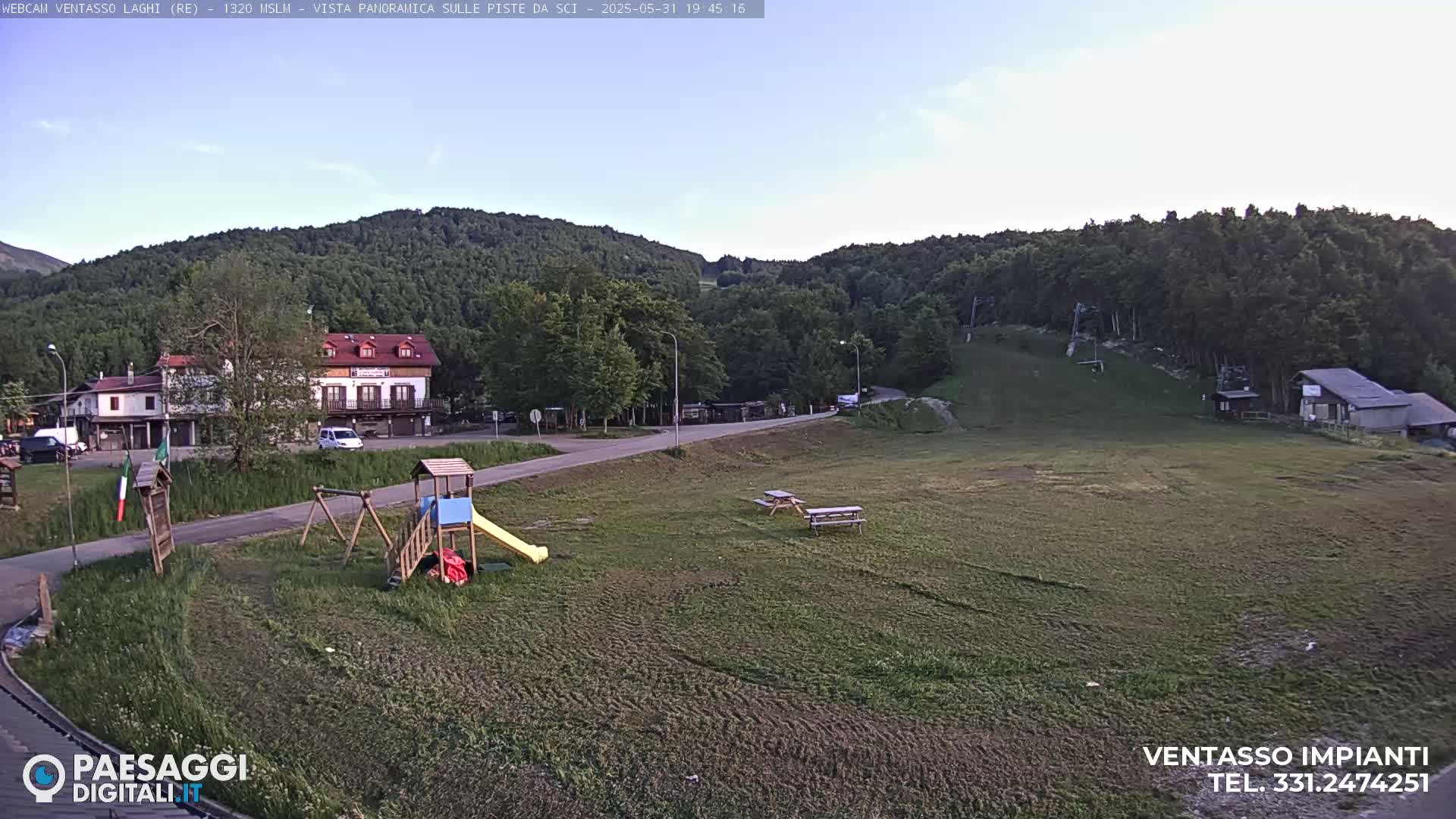 A playground and picnic tables are situated on a grassy area near buildings and a ski slope under a mostly clear sky.