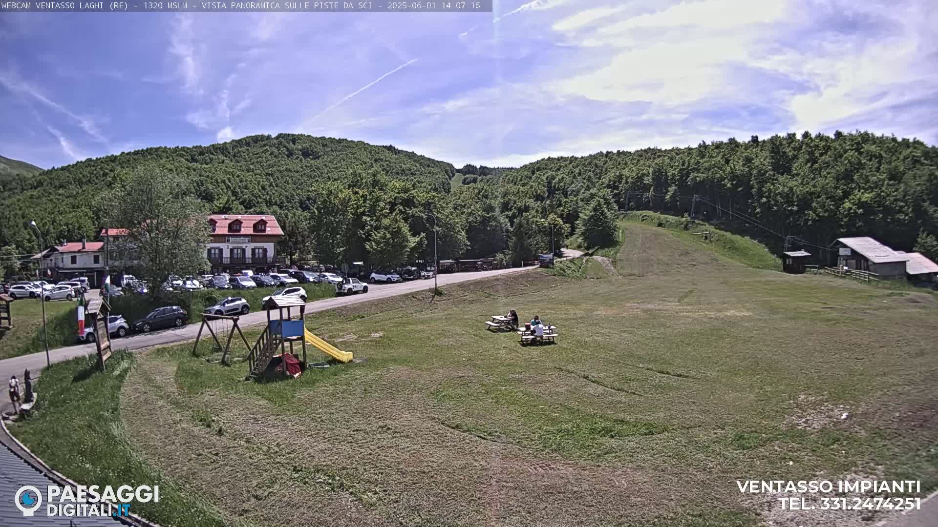 A sunny day at a ski resort shows a parking lot full of cars near a building, a playground, picnic tables with people sitting at them, and a grassy area with a ski lift in the background.