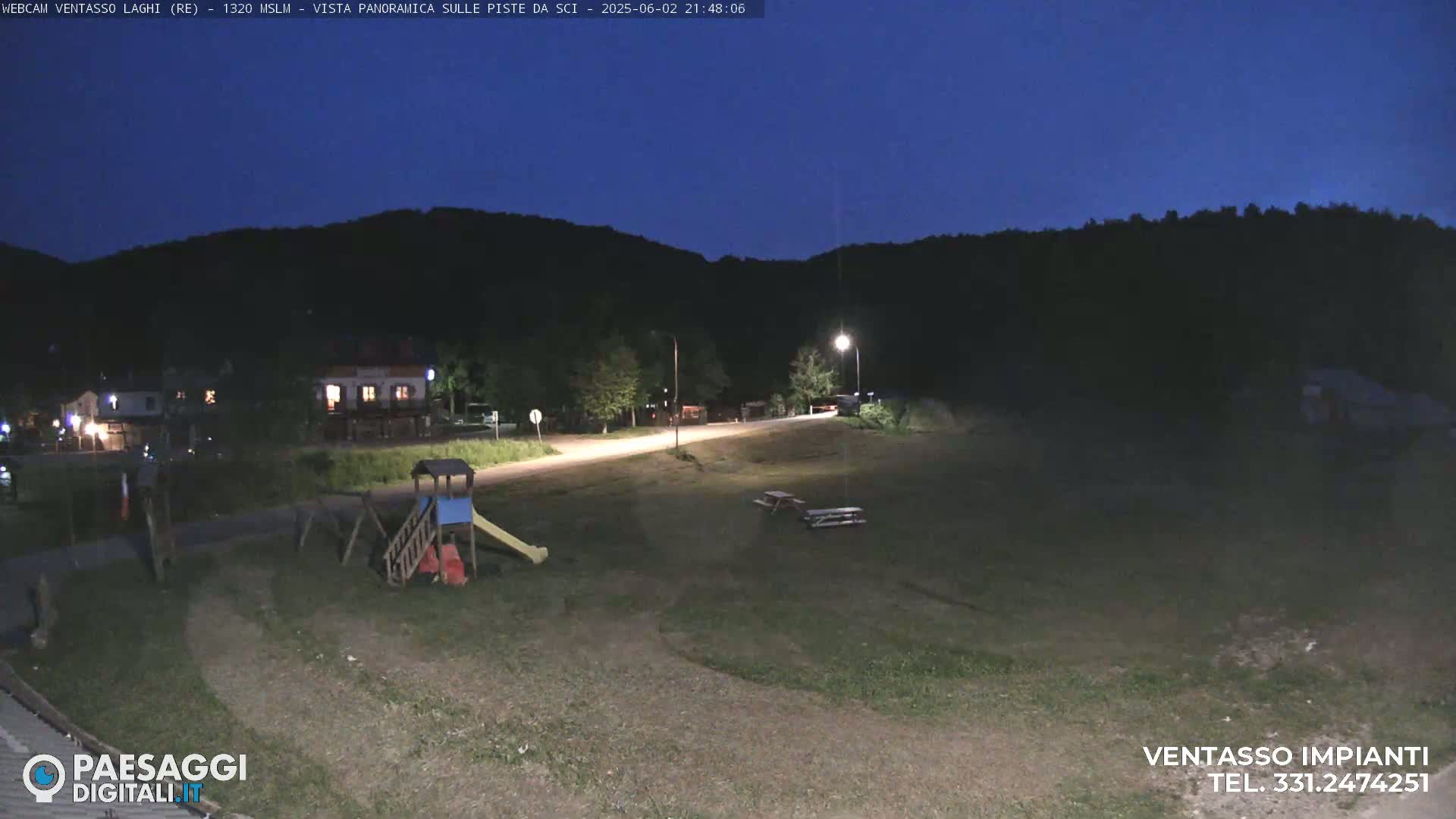 A nighttime view of a grassy area with a playground, picnic tables, and a road leading towards buildings and hills under a dark, clear sky.
