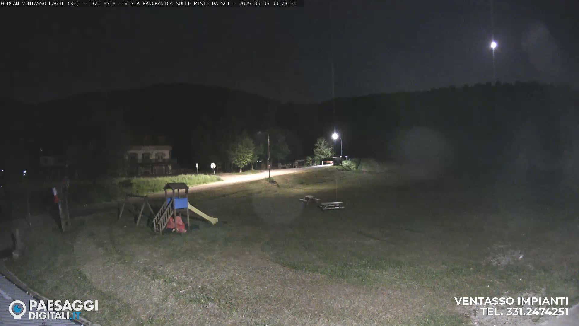 A nighttime, outdoor view shows a playground, picnic tables, and a road leading toward dark hills under a mostly clear sky with a bright light source visible in the upper-right.