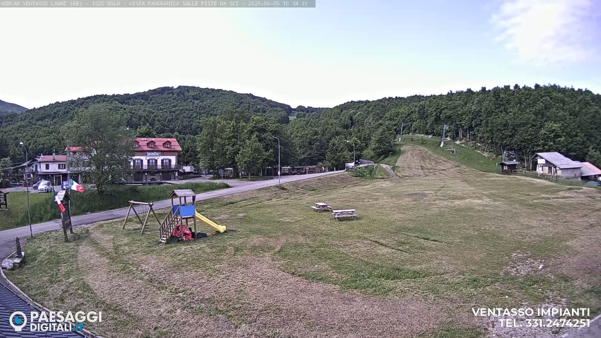 A grassy area with a playground, picnic tables, buildings, and a ski lift in the background, under a mostly clear sky.