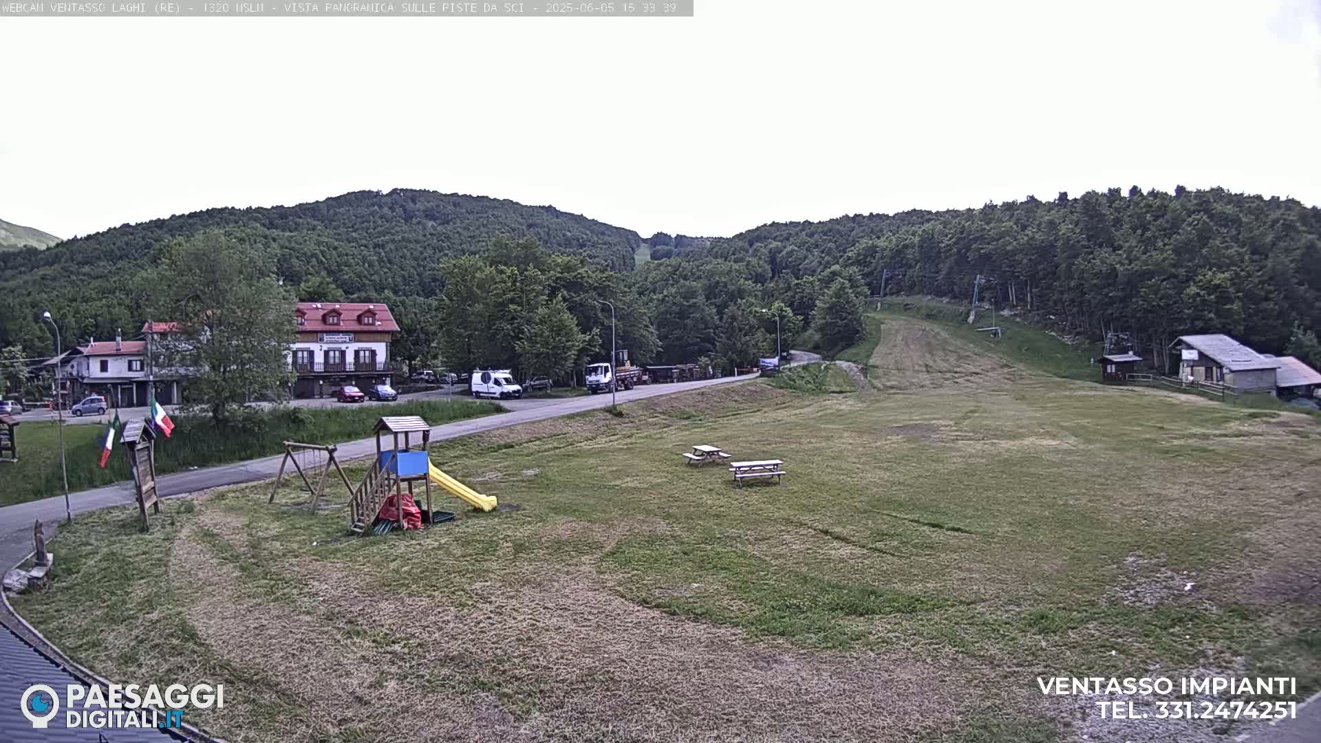 A mostly sunny view of a grassy area with a playground, picnic tables, buildings, and a ski slope in the background.