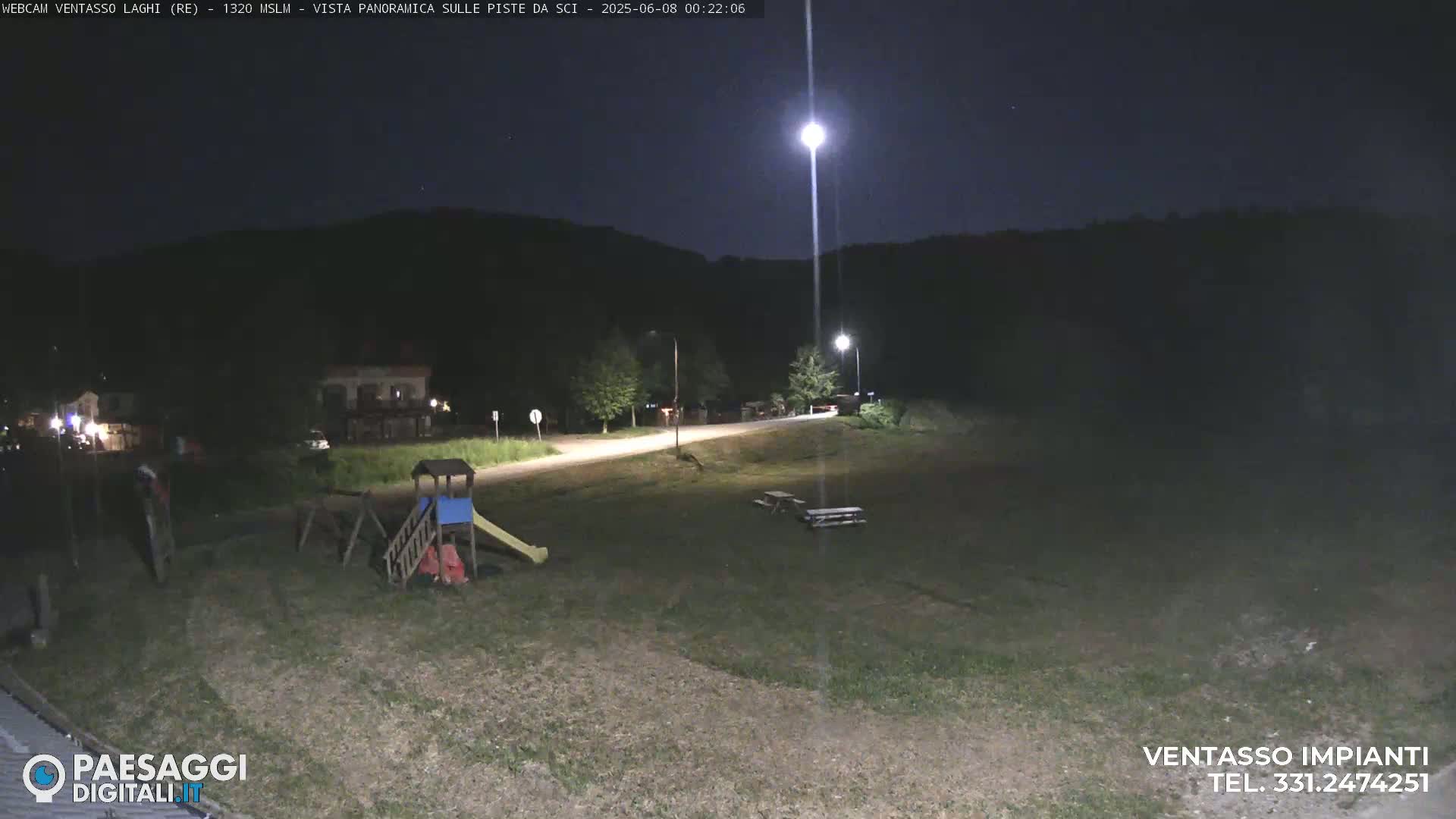 A nighttime view of a grassy area with a playground, picnic tables, and a road leading towards a hill under a bright, illuminated night sky.