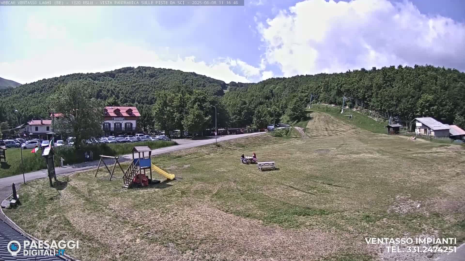 A sunny day reveals a grassy area with a playground, picnic tables, a parking lot full of cars, and buildings nestled in a mountainside setting, with a ski slope visible in the background.