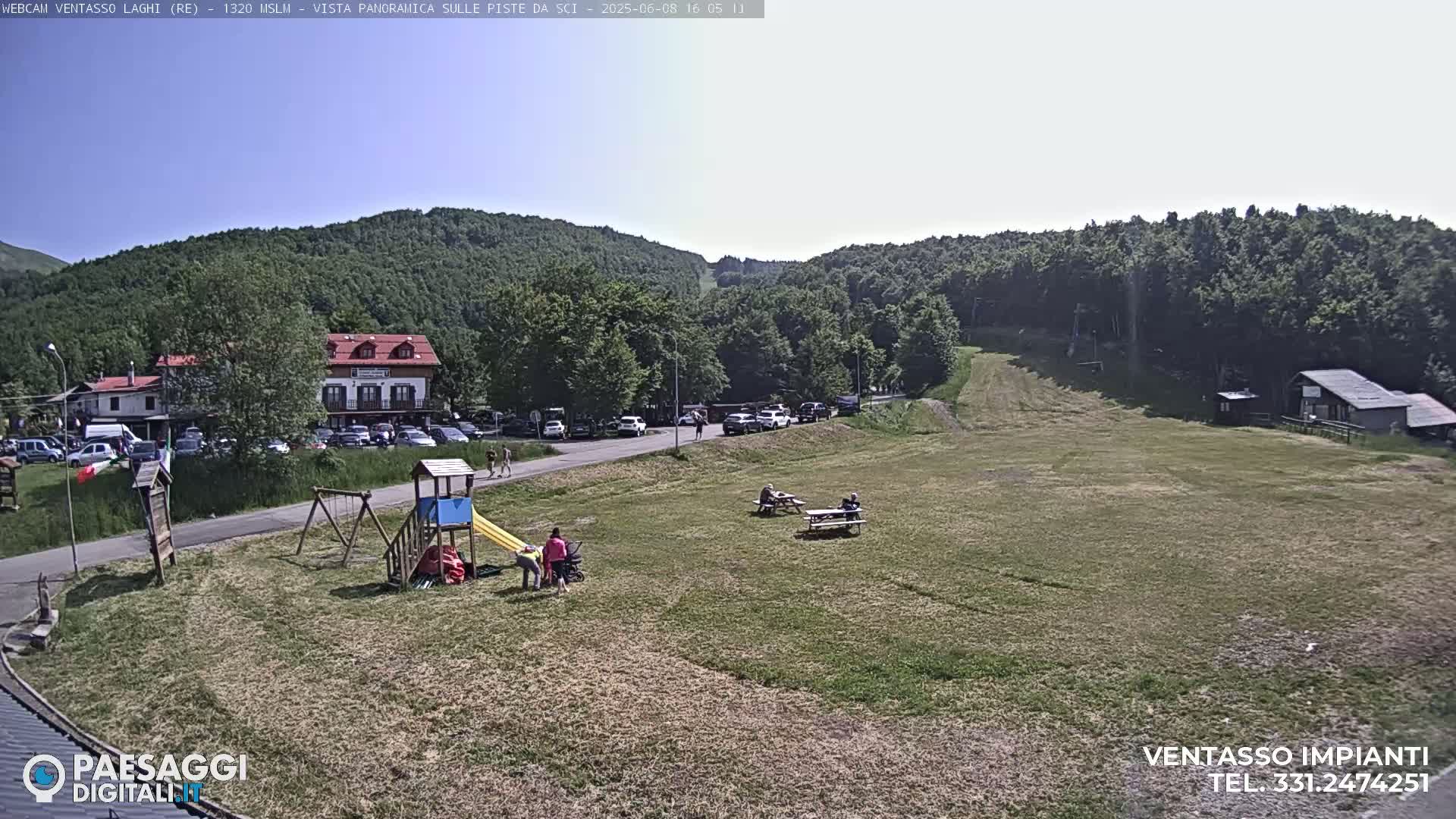 A sunny day reveals a grassy field with a playground, picnic tables, a parking lot full of cars, and buildings nestled amongst a green, tree-covered hillside.