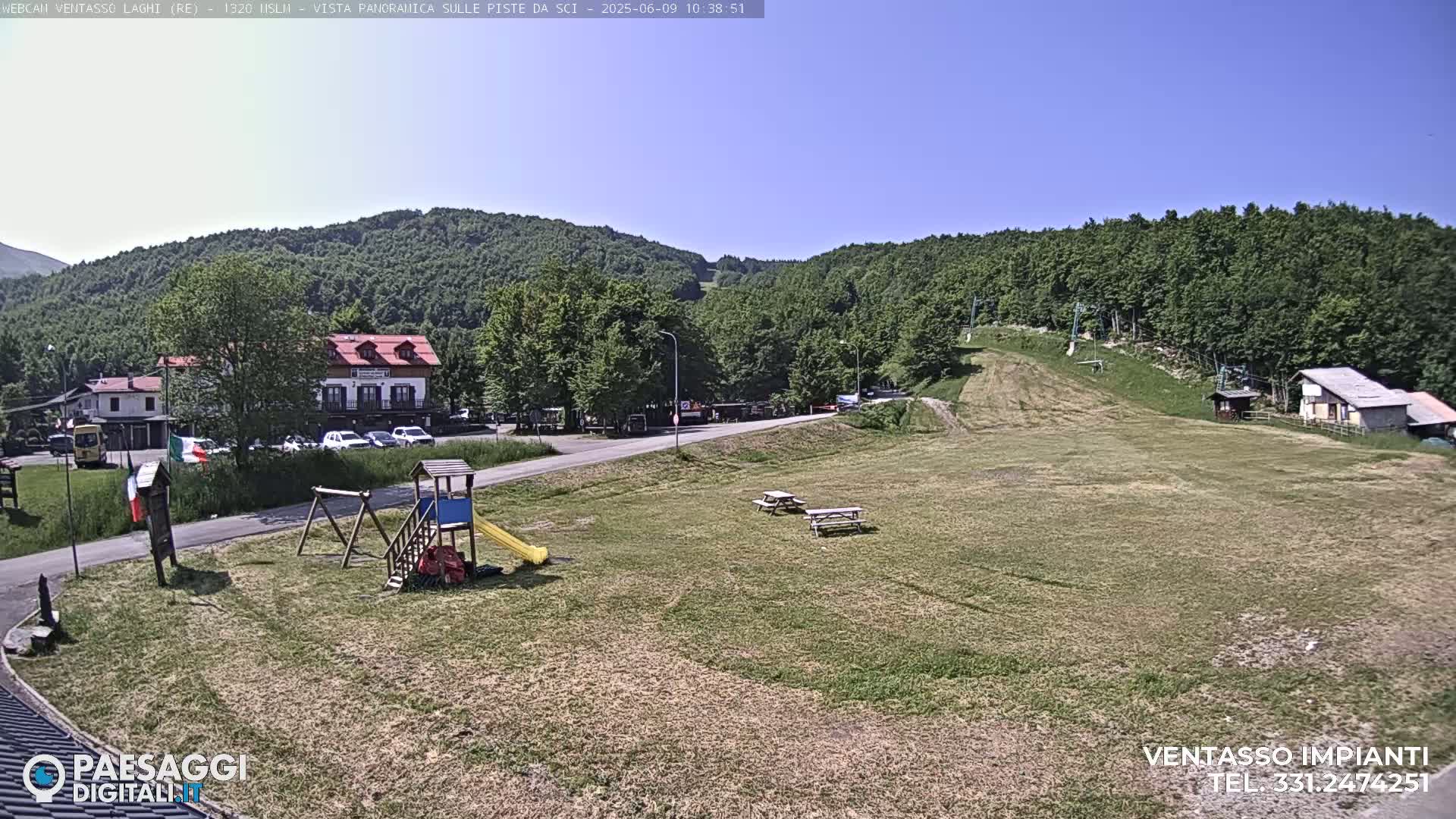 A sunny outdoor scene shows a grassy field with a playground, picnic tables, buildings, and a hill covered in lush green trees in the background.