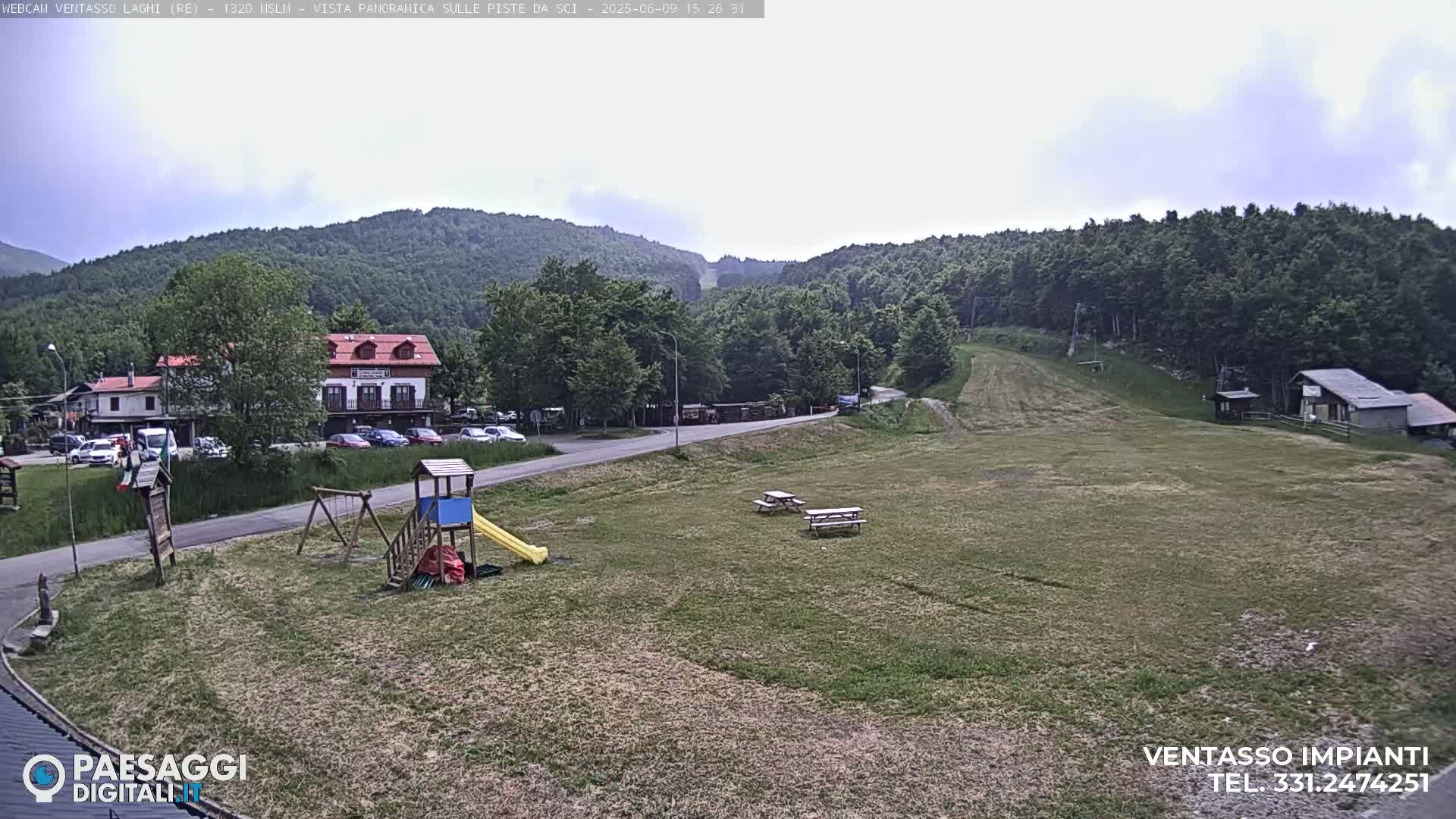 A playground and picnic tables sit on a grassy area near a road and buildings, with a mostly cloudy sky and forested hills in the background.