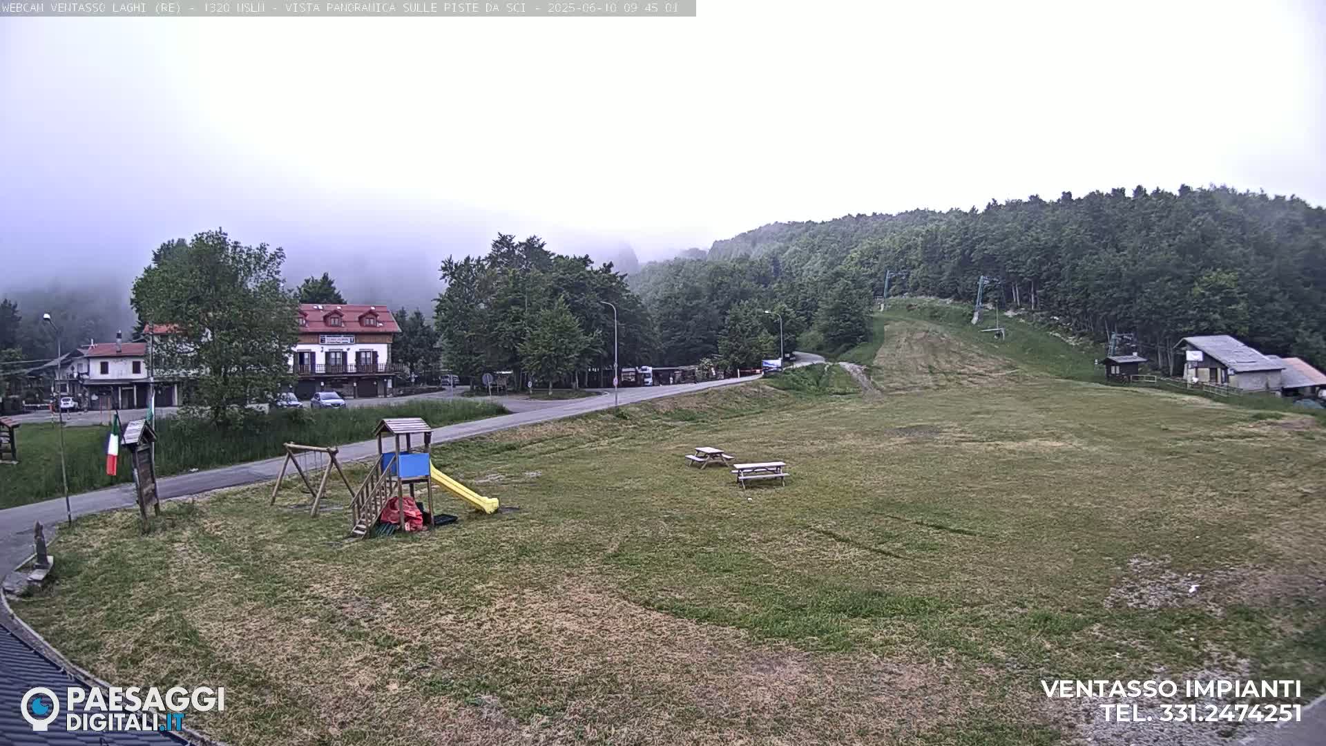 A foggy, overcast day reveals a playground, picnic tables, and buildings nestled among green hills and a ski slope.