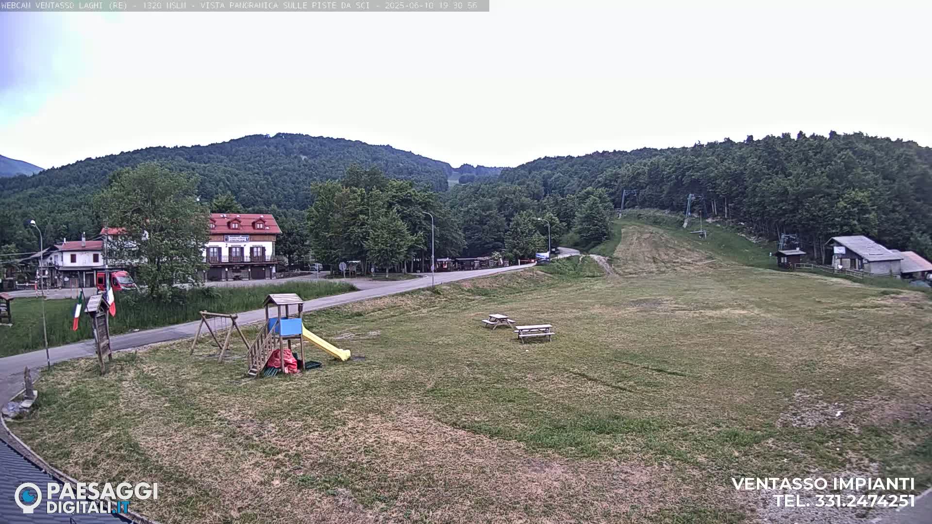 A mostly sunny view of a grassy area with a playground, picnic tables, a road, buildings, and a forested hillside in the background.