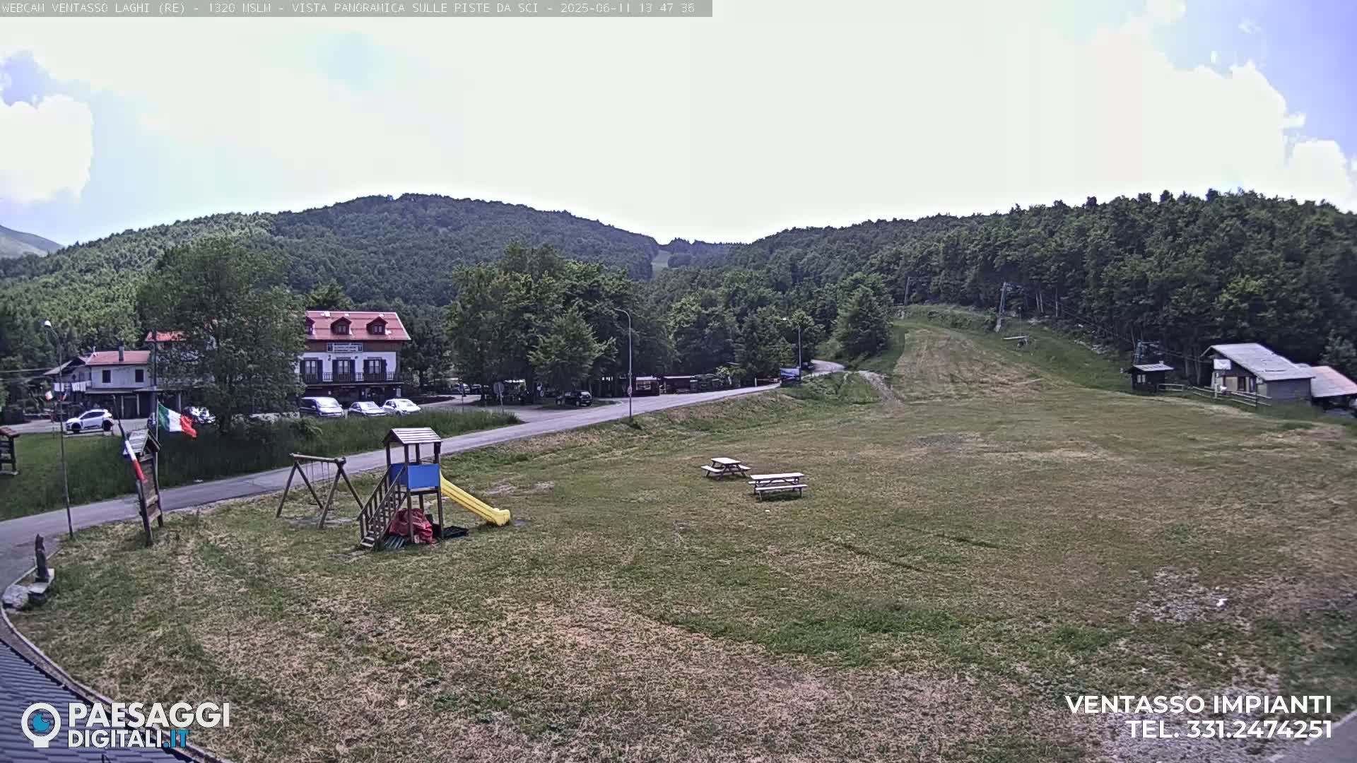 A mostly sunny view of a grassy area with a playground, picnic tables, a road, buildings, and a forested hillside in the background.