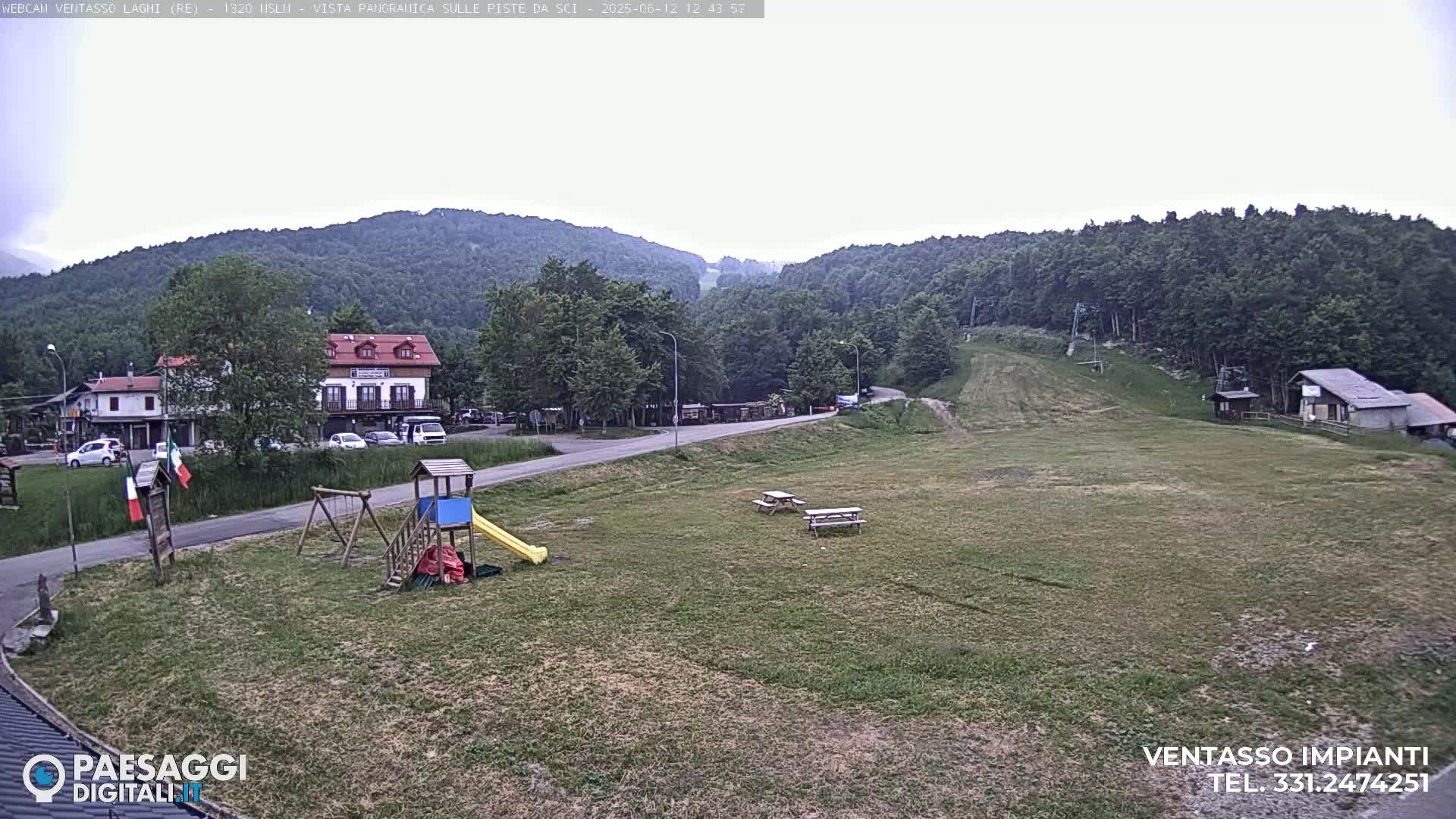 A mostly cloudy day reveals a grassy area with a playground, picnic tables, buildings, and a ski slope in the background, all nestled amongst green hills.