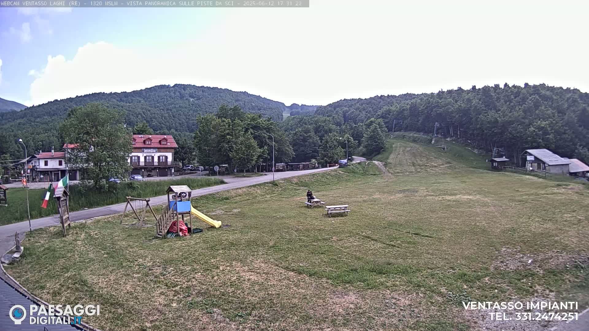 A mostly sunny view of a grassy area with a playground, picnic tables, buildings, and a forested hillside in the background.
