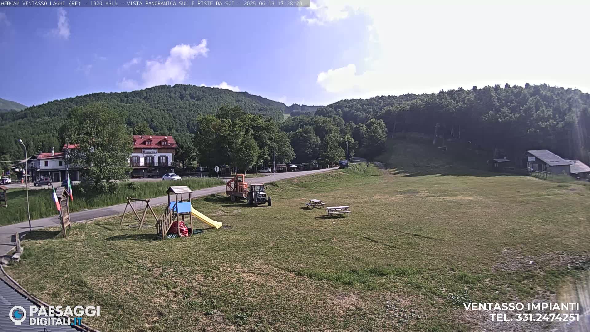 A sunny day reveals a grassy area with a playground, picnic tables, a tractor, and buildings nestled among green hills and trees.