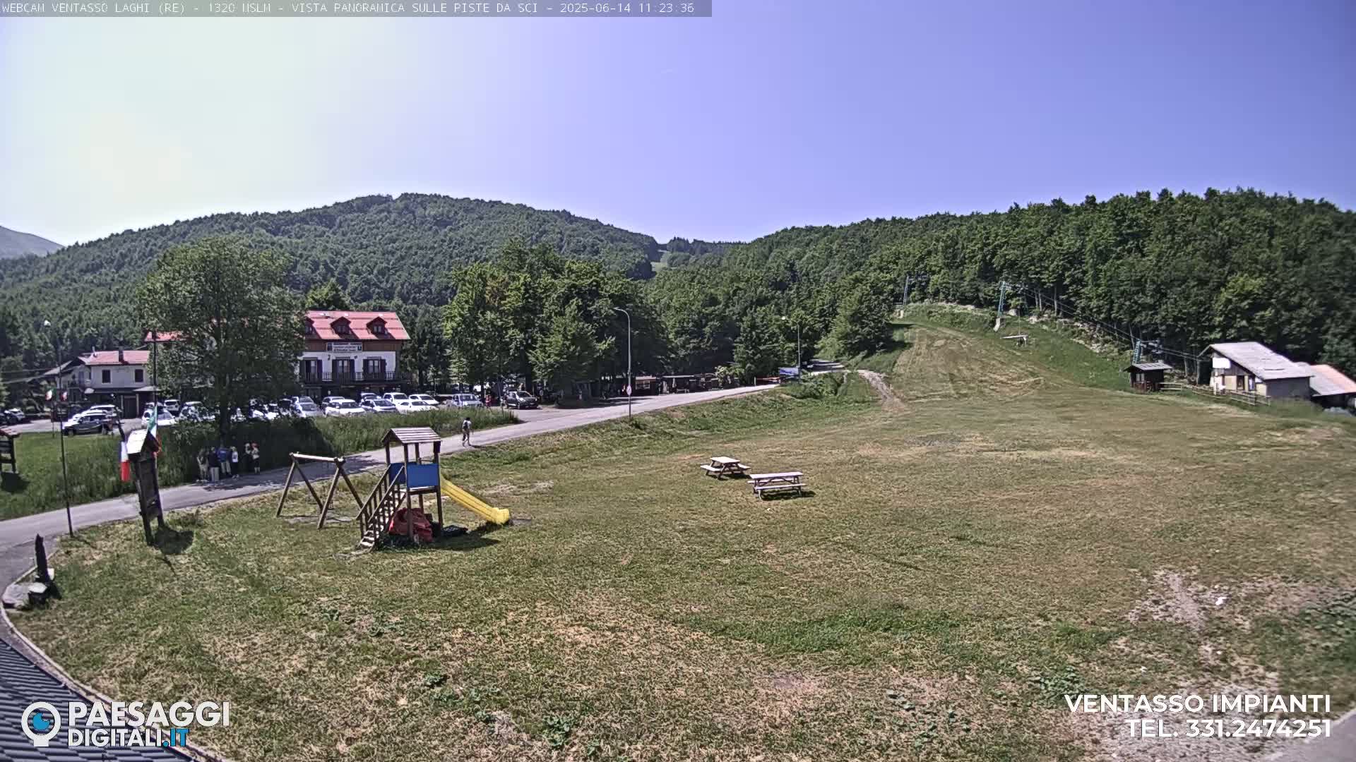 A sunny day reveals a grassy area with a playground, picnic tables, a parking lot with cars, and buildings nestled against a backdrop of green hills.