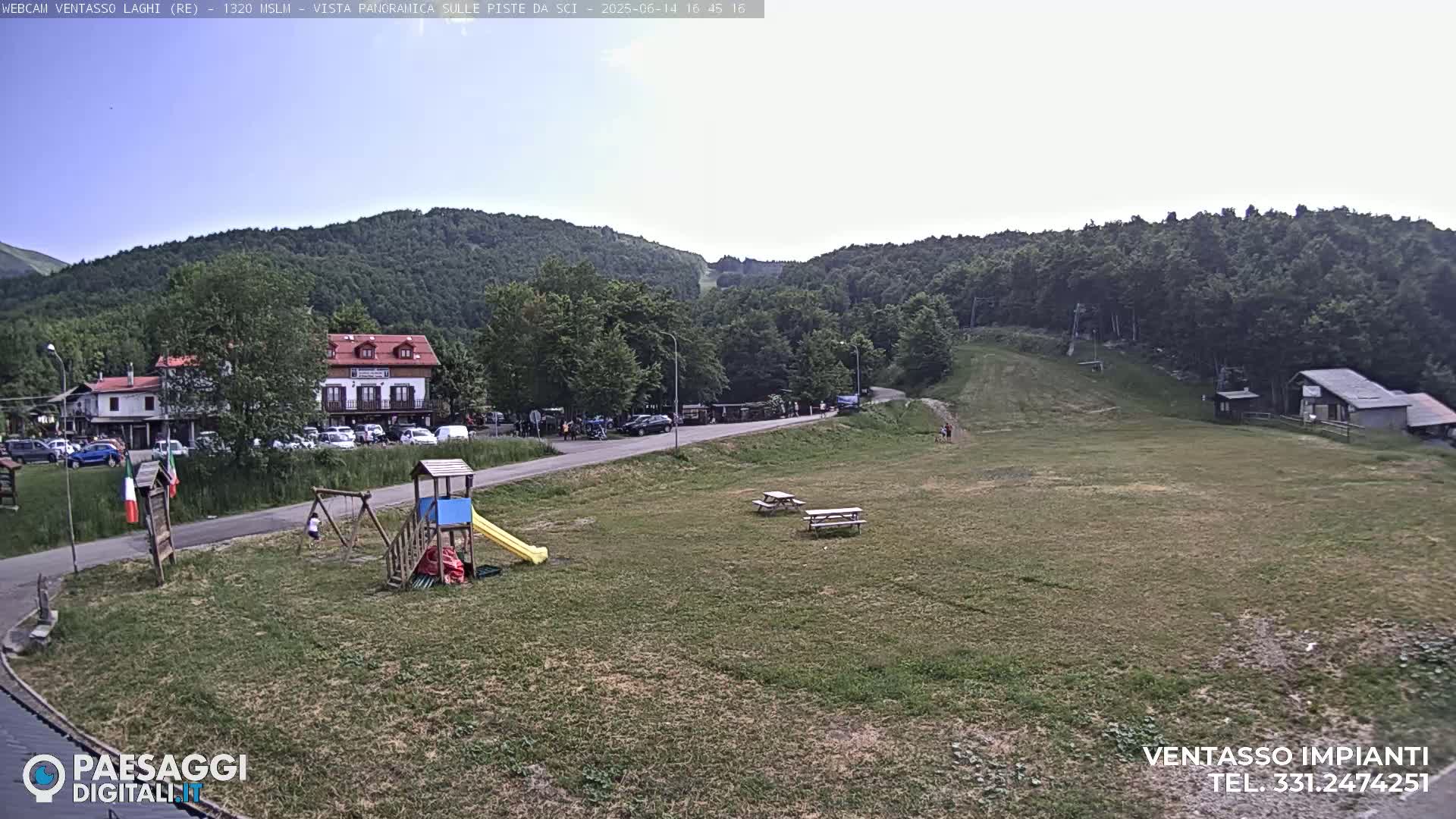 A playground and picnic tables sit on a grassy area near a road, with buildings and a ski slope visible in the background under a mostly clear sky.