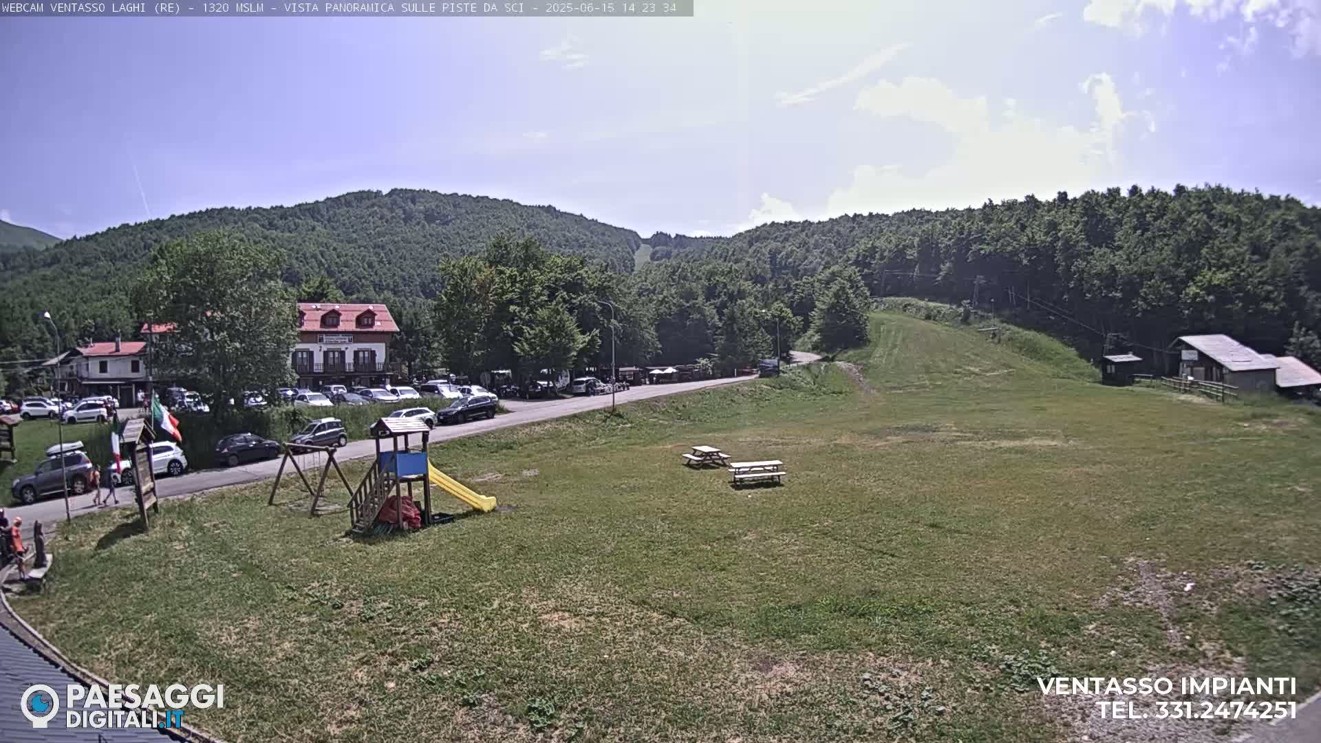 A sunny day at a mountaintop location shows a grassy area with picnic tables, a playground, a parking lot with cars, and buildings nestled among green hills.