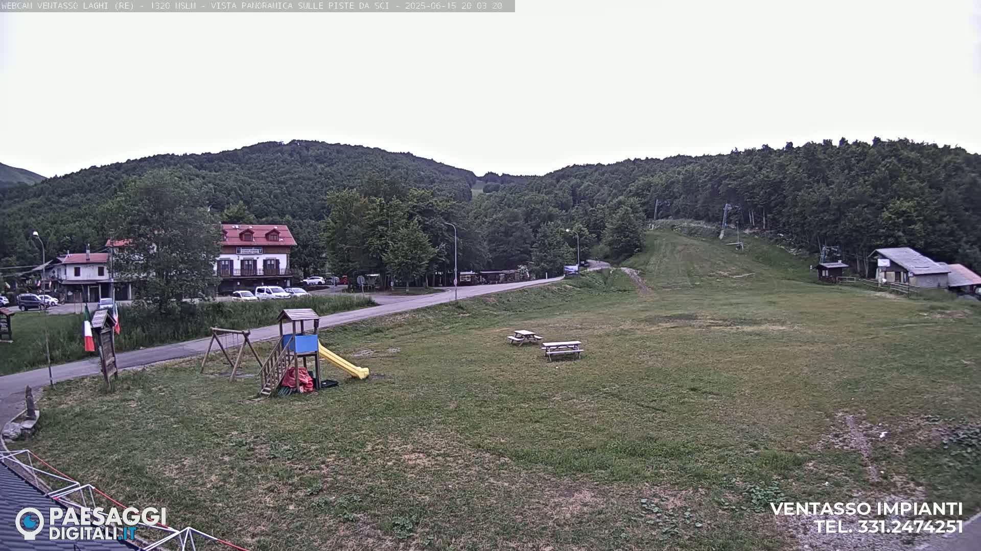A grassy area with a playground, picnic tables, and buildings nestled amongst green hills under an overcast sky.