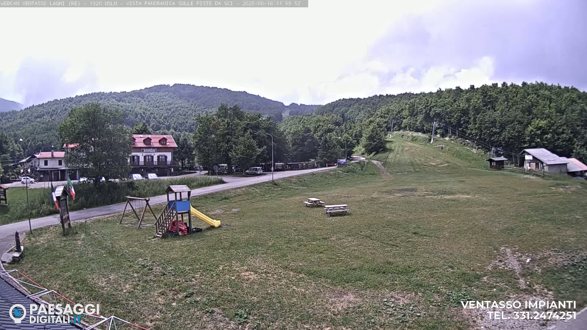 A mostly cloudy day reveals a grassy area with a playground, picnic tables, buildings, and a ski lift against a backdrop of green, tree-covered hills.