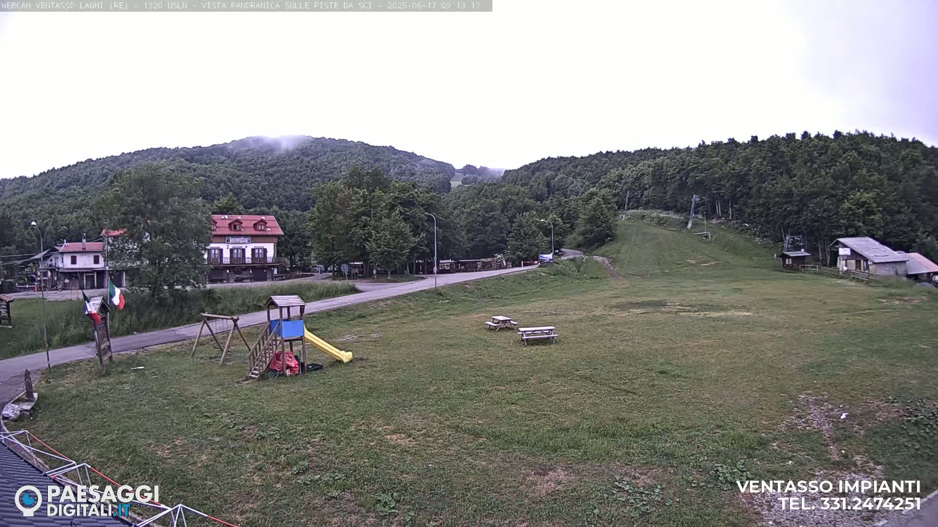 A playground and picnic tables sit on a grassy field, overlooking a road, buildings, and a forested hillside partially shrouded in fog.