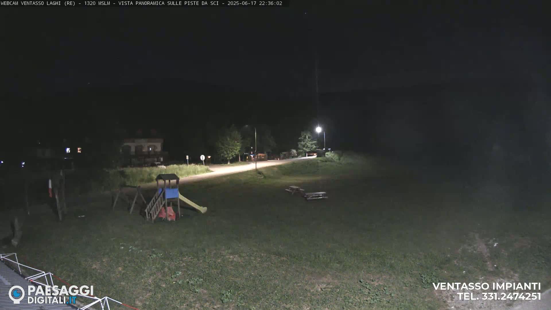 A nighttime view of a grassy area with a playground and picnic tables near a road, illuminated by streetlights.