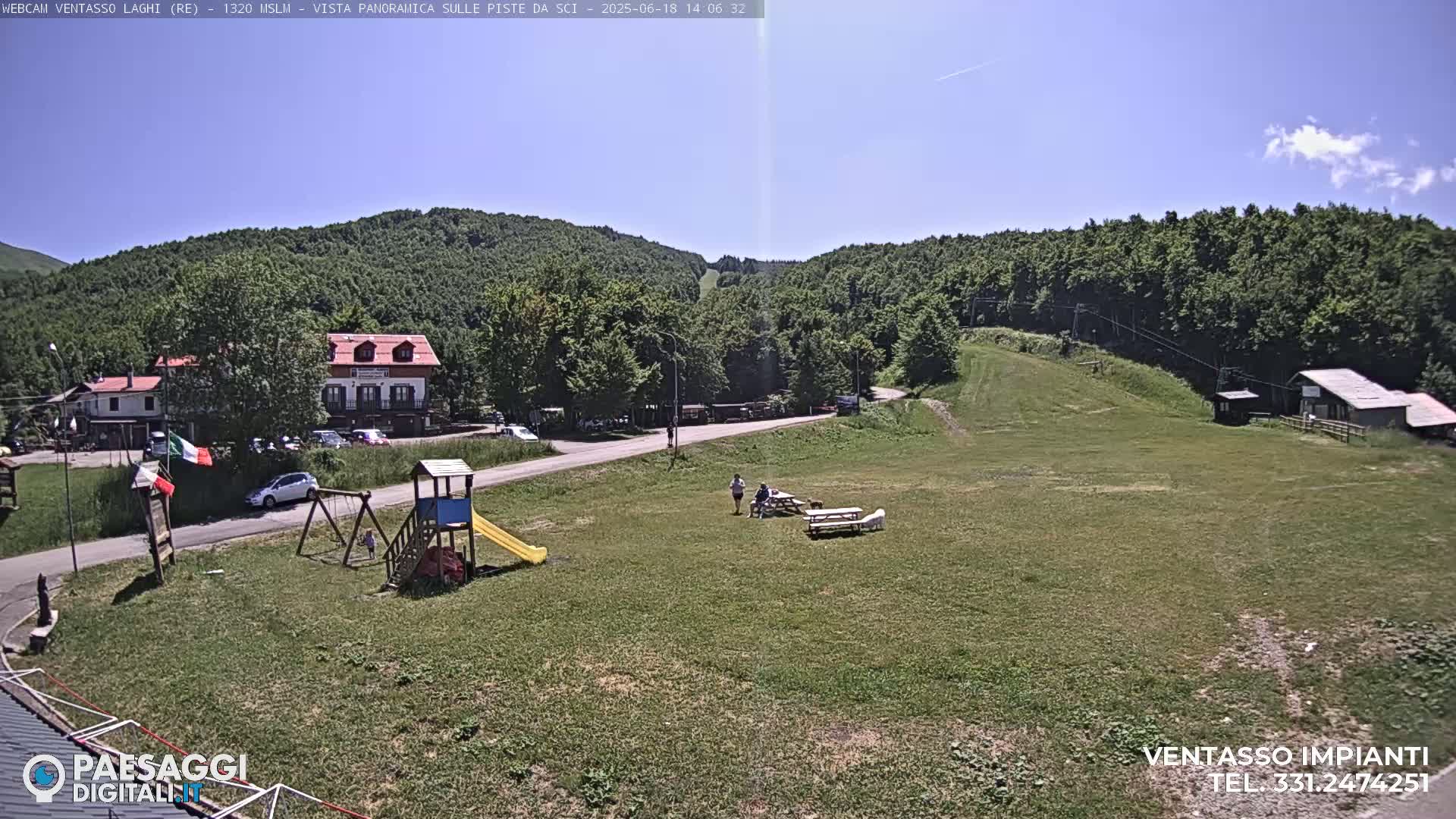 A sunny day at a mountain resort shows a grassy area with a playground, a few people, buildings, and a ski lift in the background.