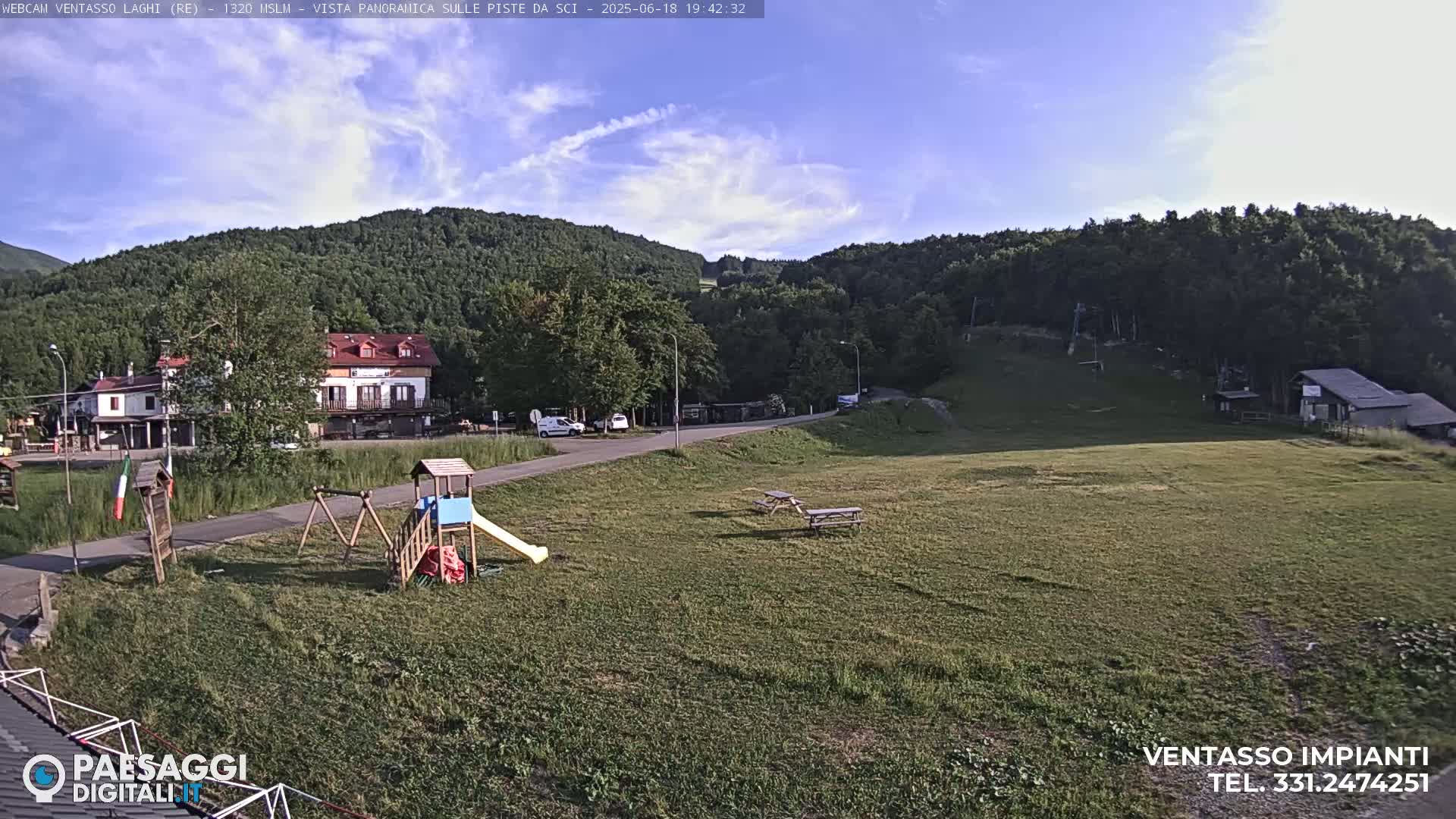 A mostly sunny view of a grassy area with a playground, picnic tables, a few buildings, and a forested hillside in the background.