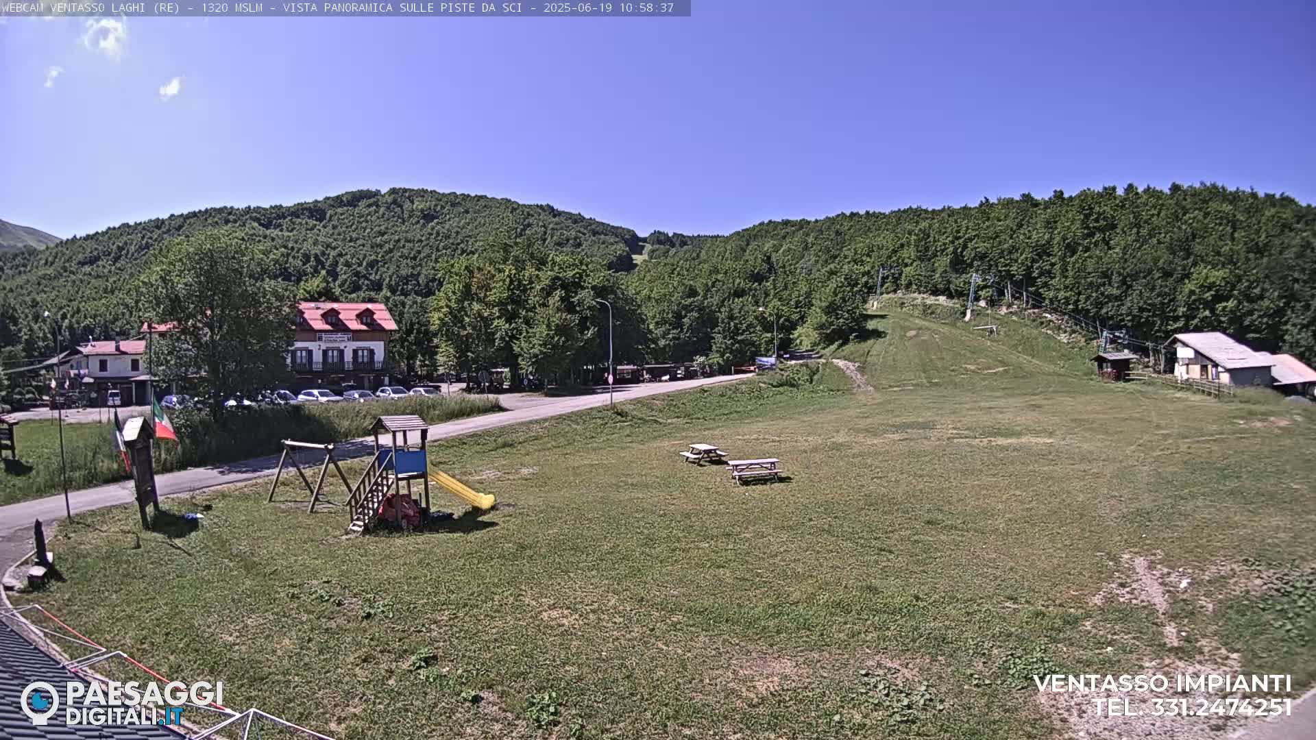 A sunny view of a grassy area with a playground, picnic tables, buildings, and a ski lift in the background, surrounded by green hills.