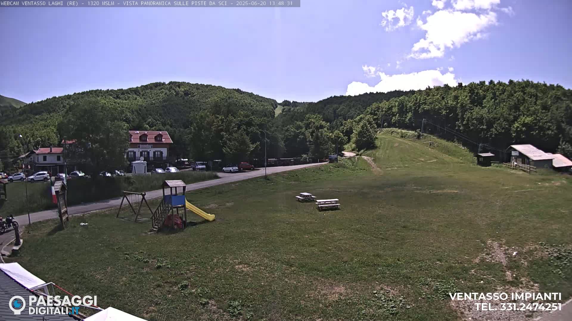 A sunny day shows a grassy area with a playground, picnic tables, a small building, and a parking lot near a building and a hillside covered in green trees.