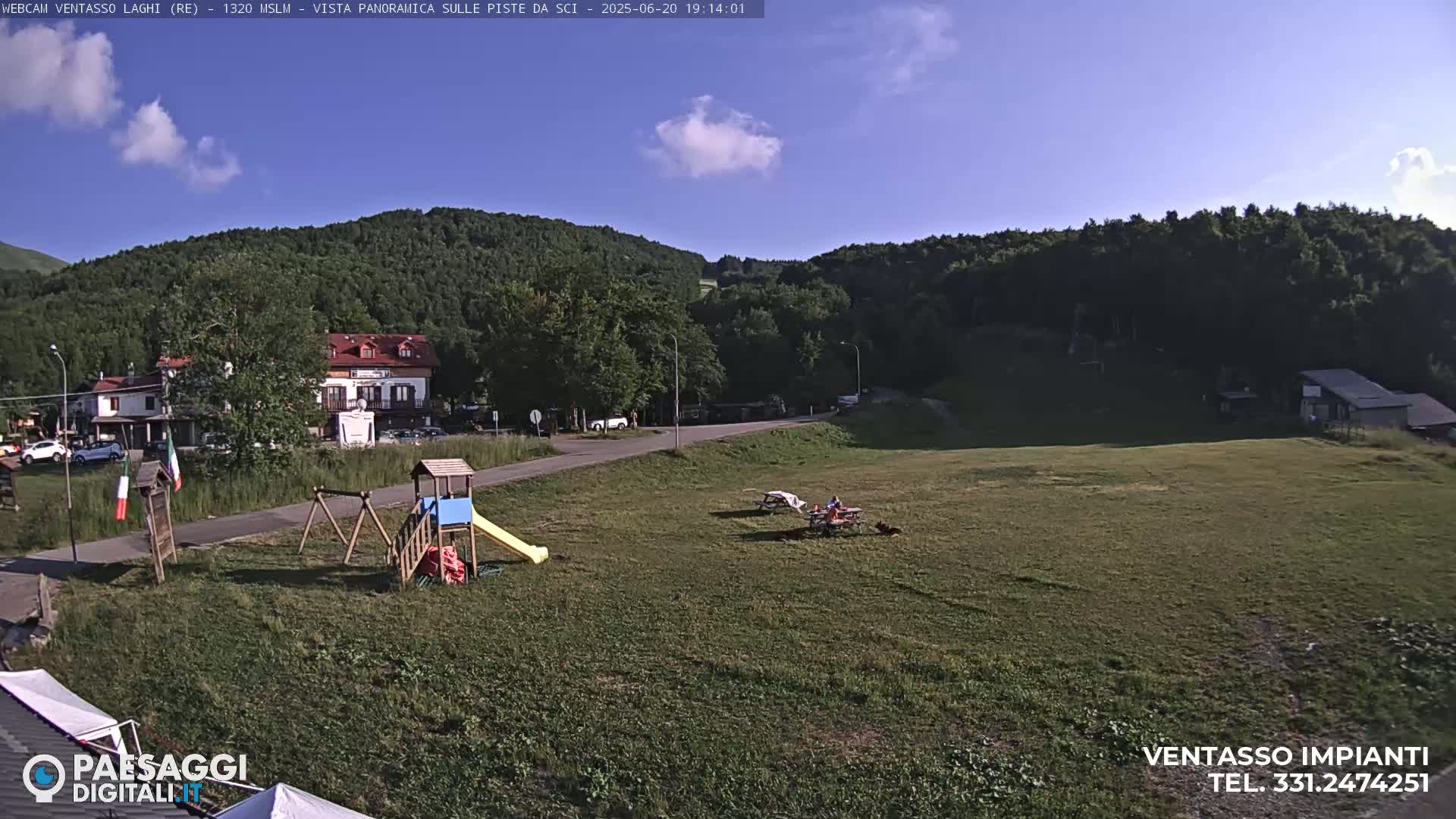 A grassy field with a playground, picnic tables, and a small building is situated below a tree-covered mountain under a mostly clear blue sky.