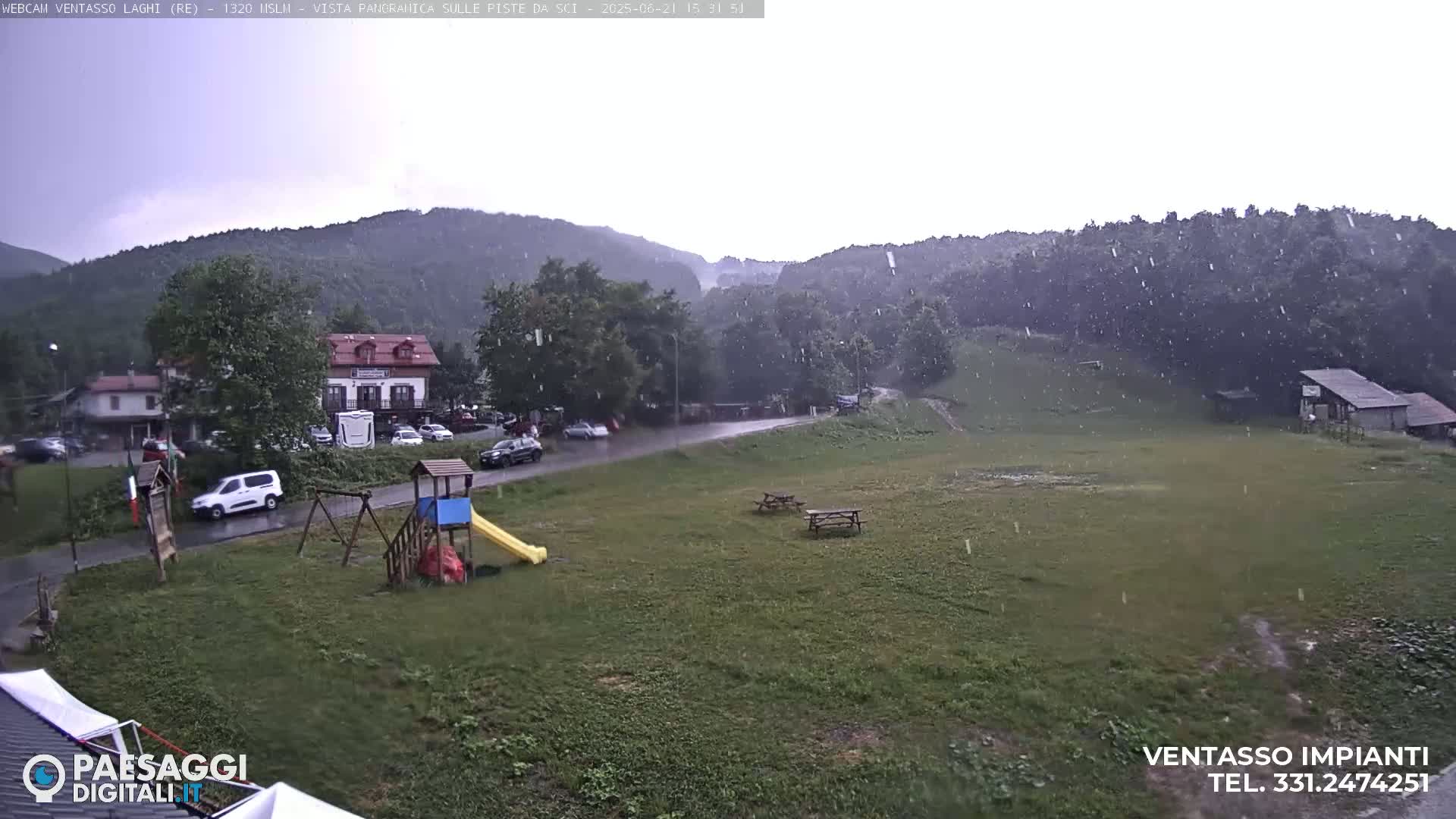 A grassy field with playground equipment and picnic tables is seen during a light rain shower in front of a mountainous landscape with buildings in the background.