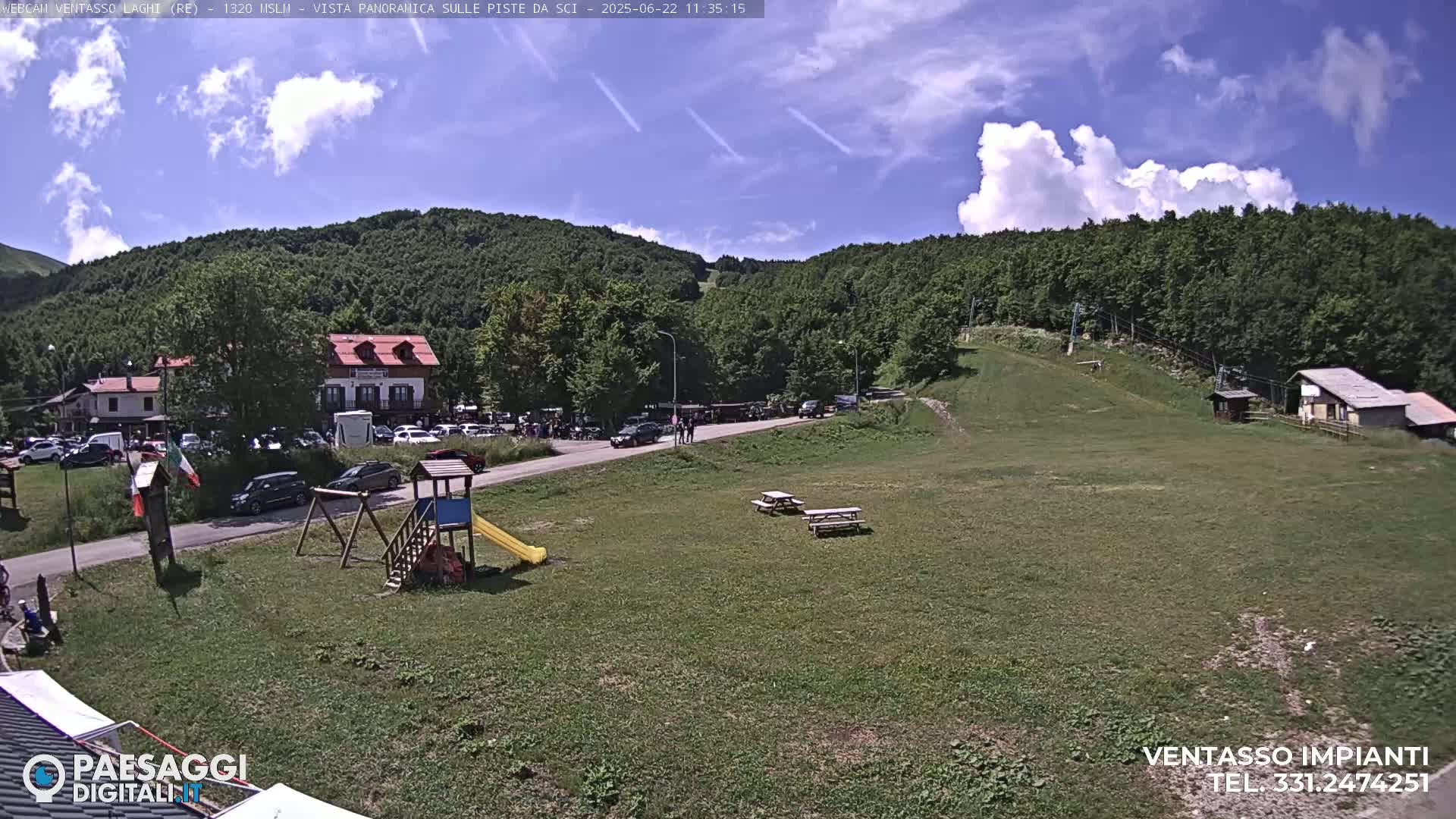 A sunny day with some clouds in a mountainous area shows a parking lot full of cars near buildings and a grassy field with picnic tables and a playground.