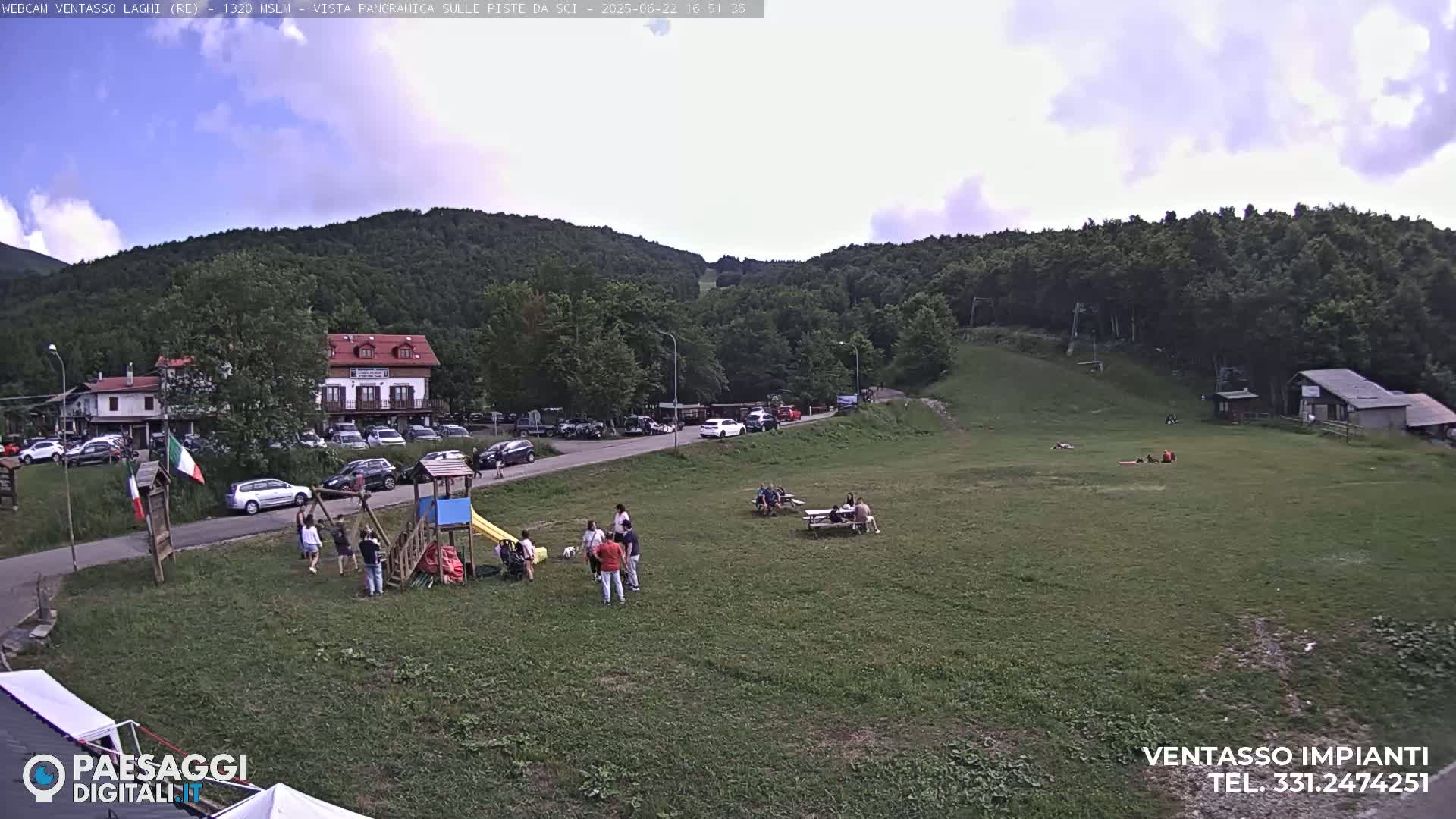 A grassy area with a playground, people relaxing at picnic tables, and a parking lot is nestled beside a hillside covered in lush green trees under a partly cloudy sky.