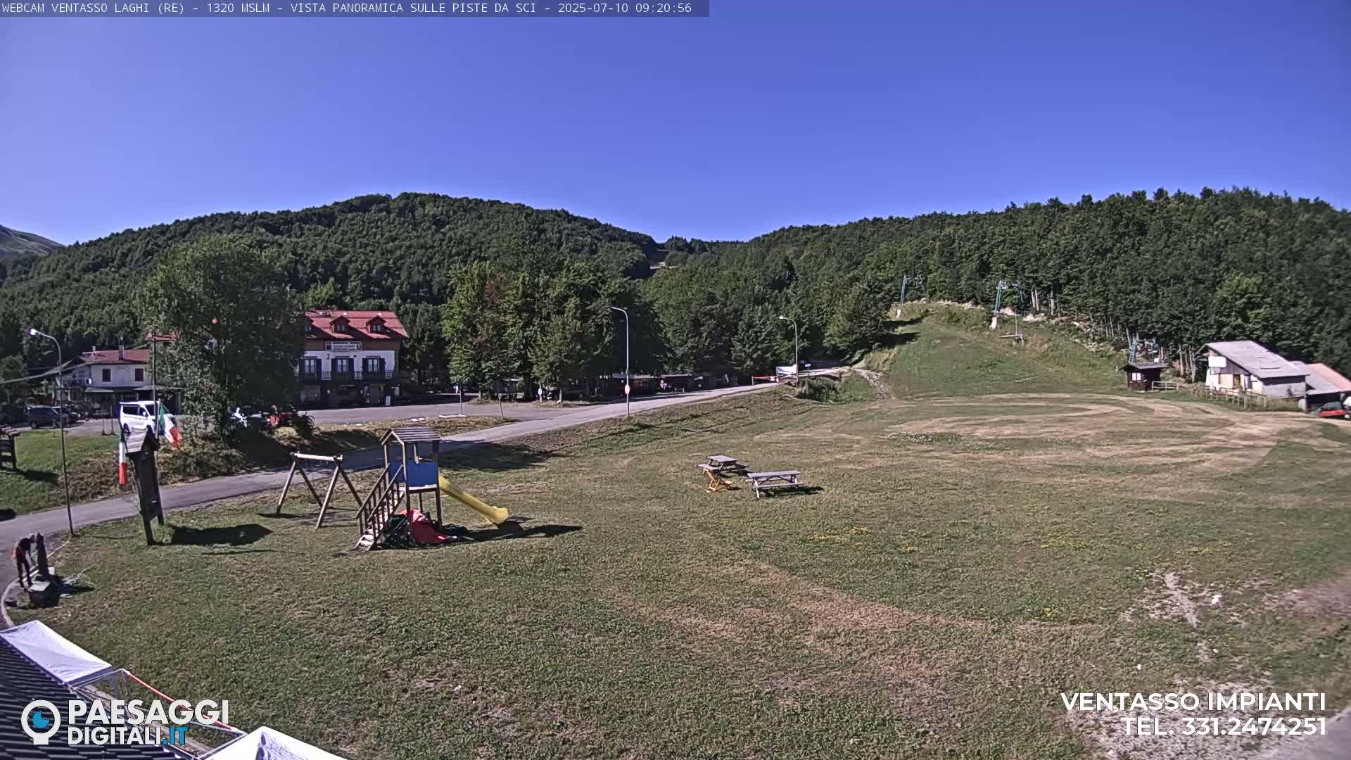 A sunny day reveals a grassy area with a playground, picnic tables, buildings, and a forested hillside in the background.