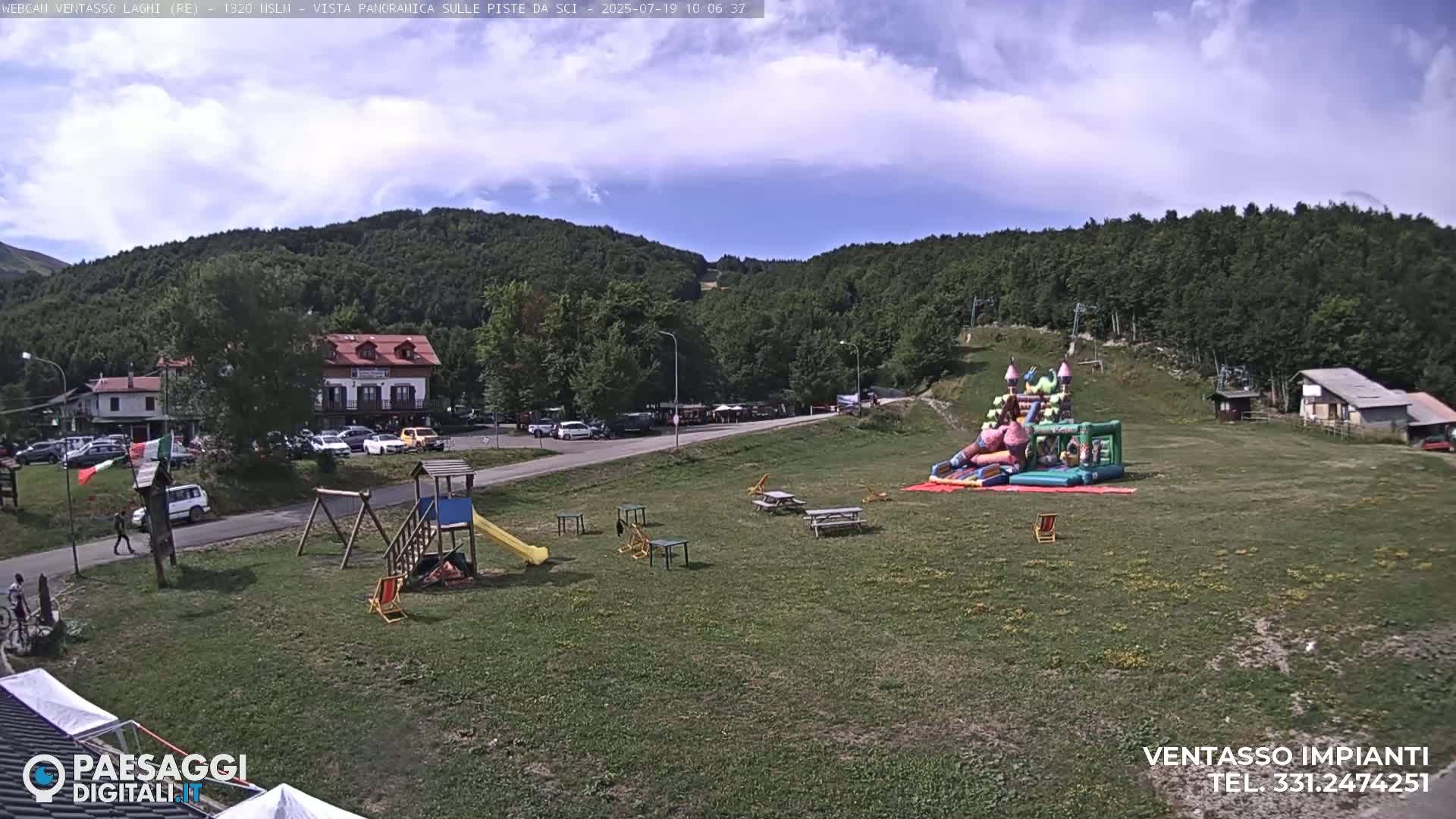 A grassy area with a playground, picnic tables, and an inflatable bounce house is nestled between a parking lot, buildings, and a hillside covered in lush green trees under a partly cloudy sky.