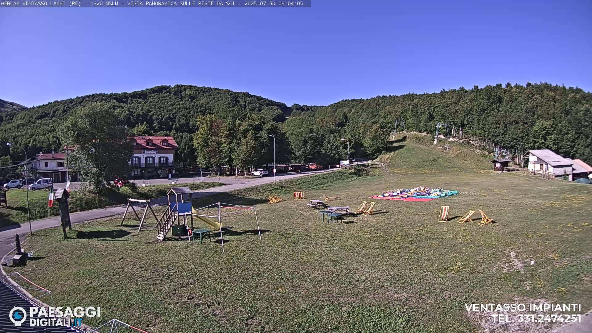 A grassy area with a playground, picnic tables, and lounge chairs sits below a hillside covered in green trees under a clear blue sky.