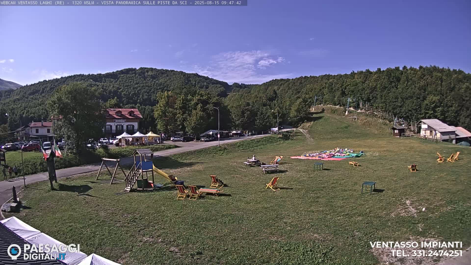 A sunny day reveals a grassy area with a playground, picnic tables, and lounge chairs, situated below a forested hillside and near buildings and a parking lot.