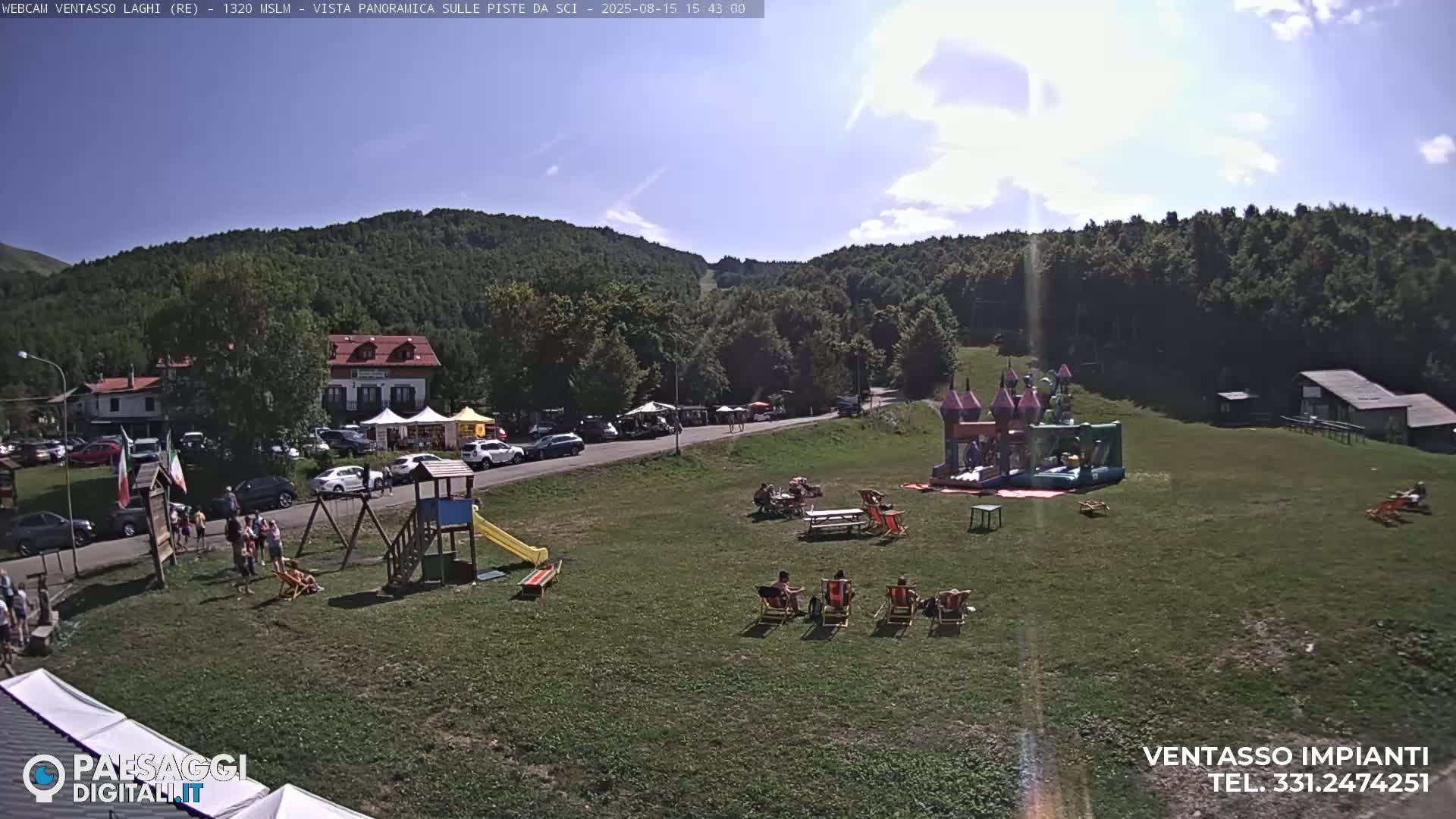 A sunny day reveals a grassy area with a playground, picnic tables, and lounge chairs, situated below a forested hillside and near buildings and a parking lot.
