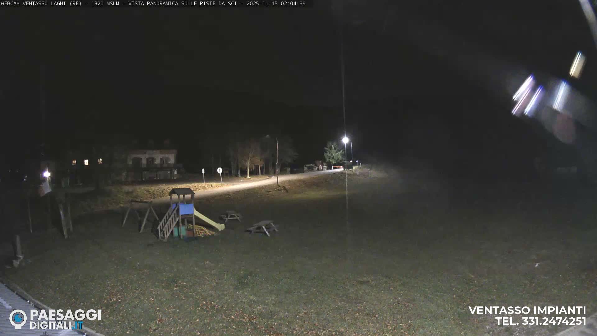 A clear night reveals a brightly lit playground and picnic tables on a grassy field, with a winding road, streetlights, and buildings in the background.