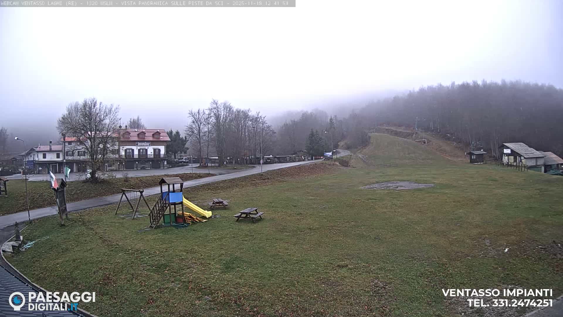 A clear night reveals a brightly lit playground and picnic tables on a grassy field, with a winding road, streetlights, and buildings in the background.