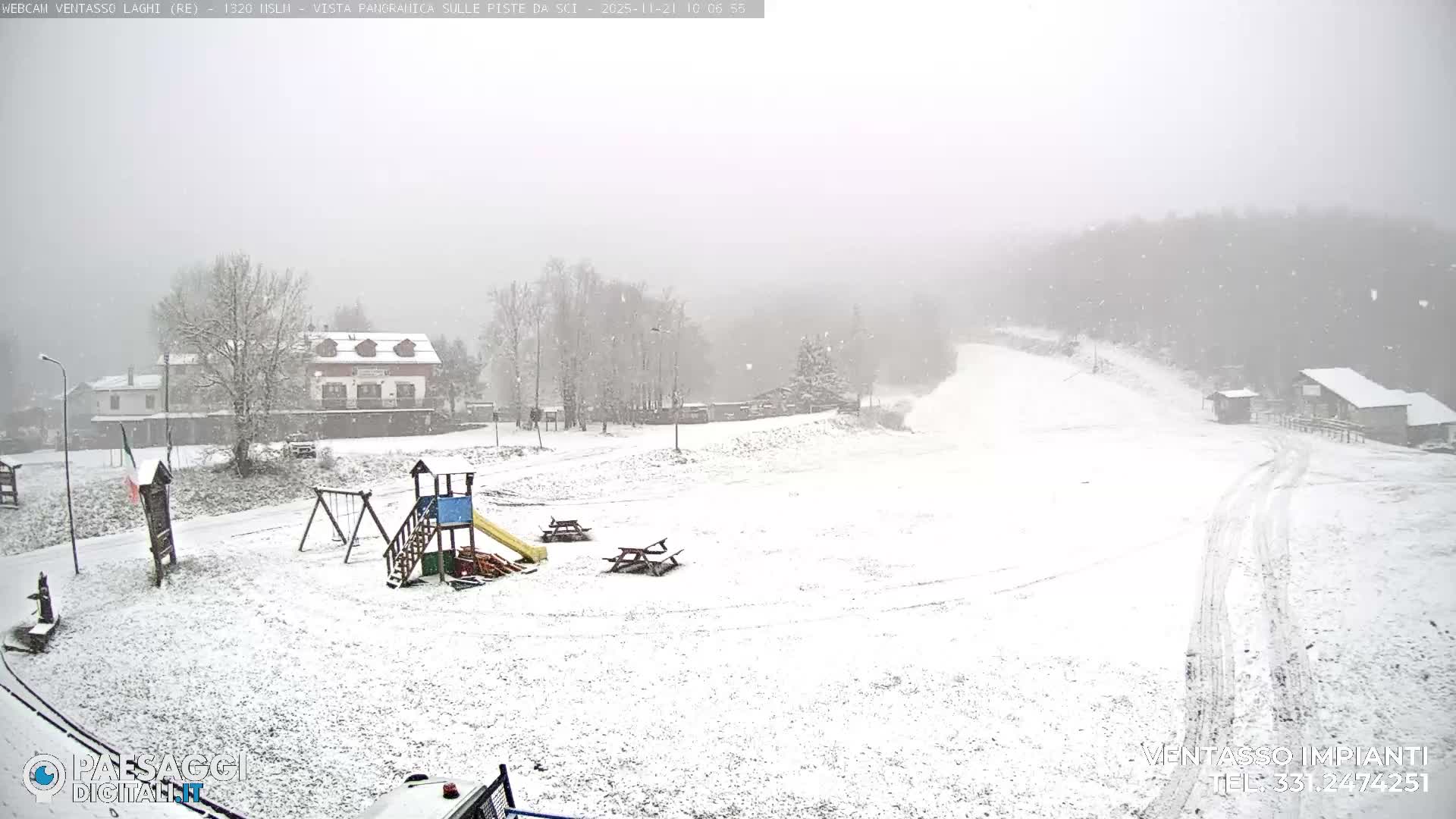 A snow-covered village at the base of a ski slope, featuring buildings, trees, a playground, and picnic tables, is seen under active heavy snowfall and hazy conditions.