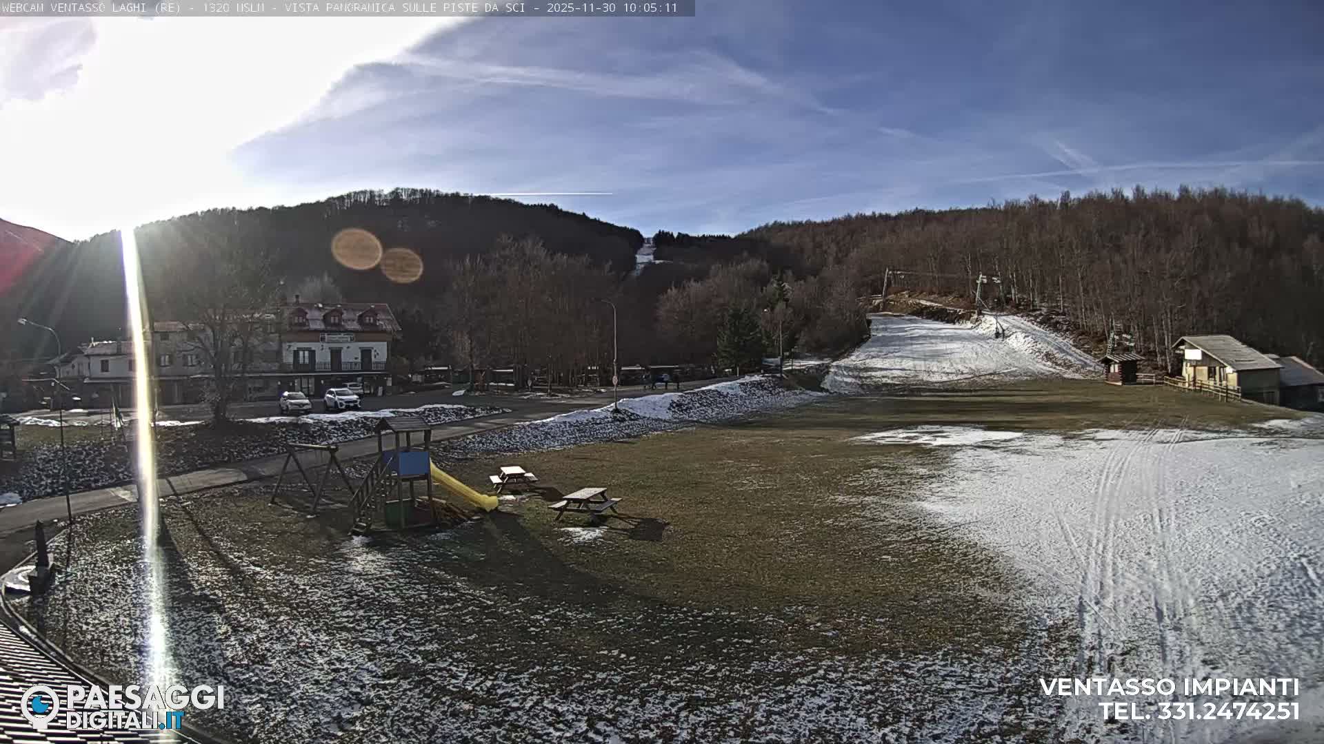 Under a bright, sunny sky with wispy clouds, a partially snow-dusted ski resort base features a playground, picnic tables, and buildings, with a ski slope and lift visible on the surrounding forested mountains.
