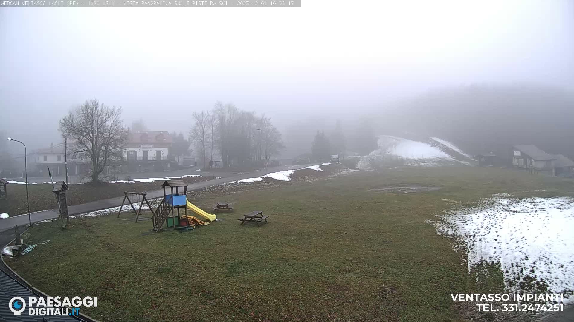 A foggy outdoor scene reveals a green field with patches of snow, a children's playground, and picnic tables in the foreground, leading to buildings and bare trees partially hidden by dense mist in the midground and background.