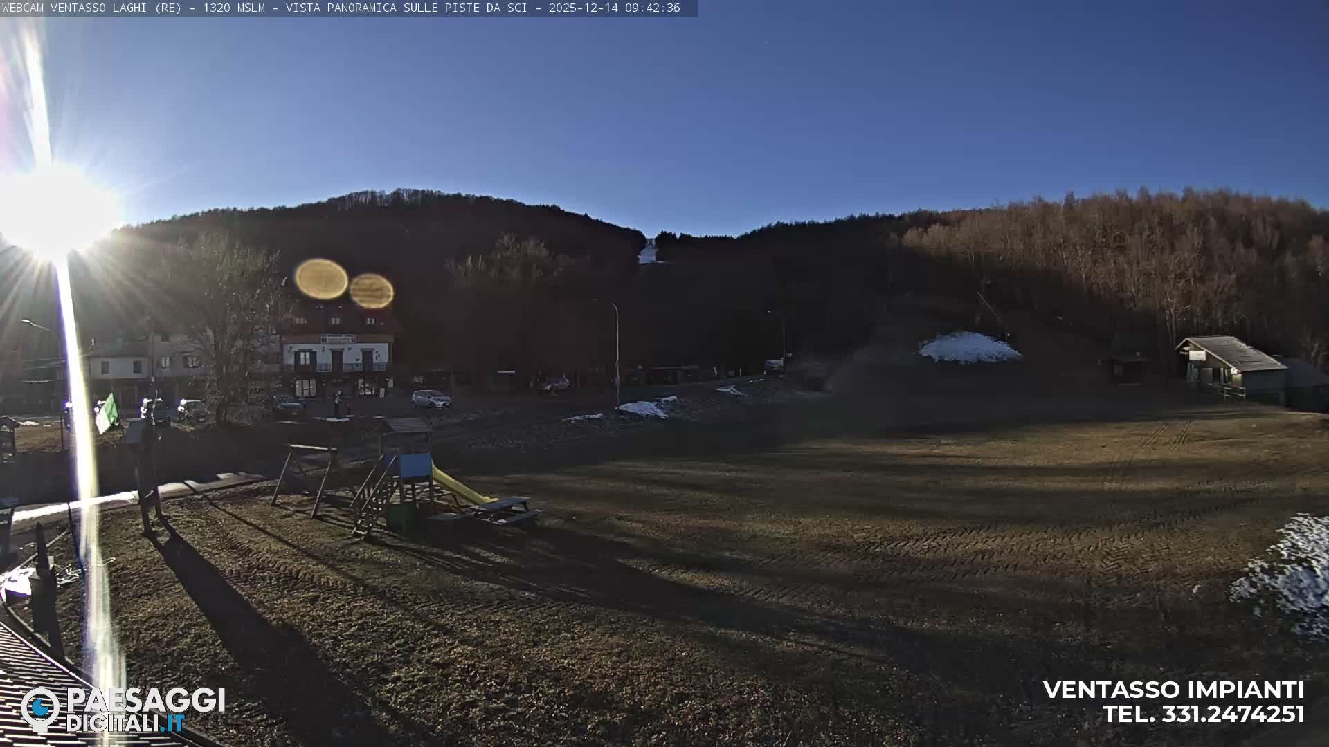 A foggy outdoor scene reveals a green field with patches of snow, a children's playground, and picnic tables in the foreground, leading to buildings and bare trees partially hidden by dense mist in the midground and background.
