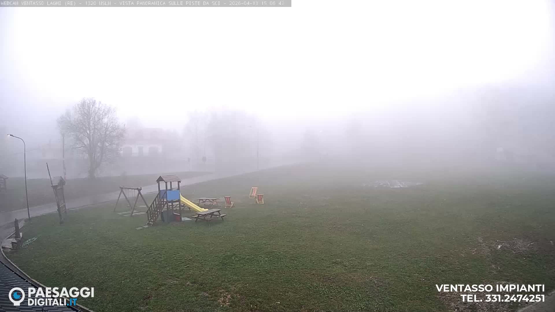 A foggy outdoor scene reveals a green field with patches of snow, a children's playground, and picnic tables in the foreground, leading to buildings and bare trees partially hidden by dense mist in the midground and background.