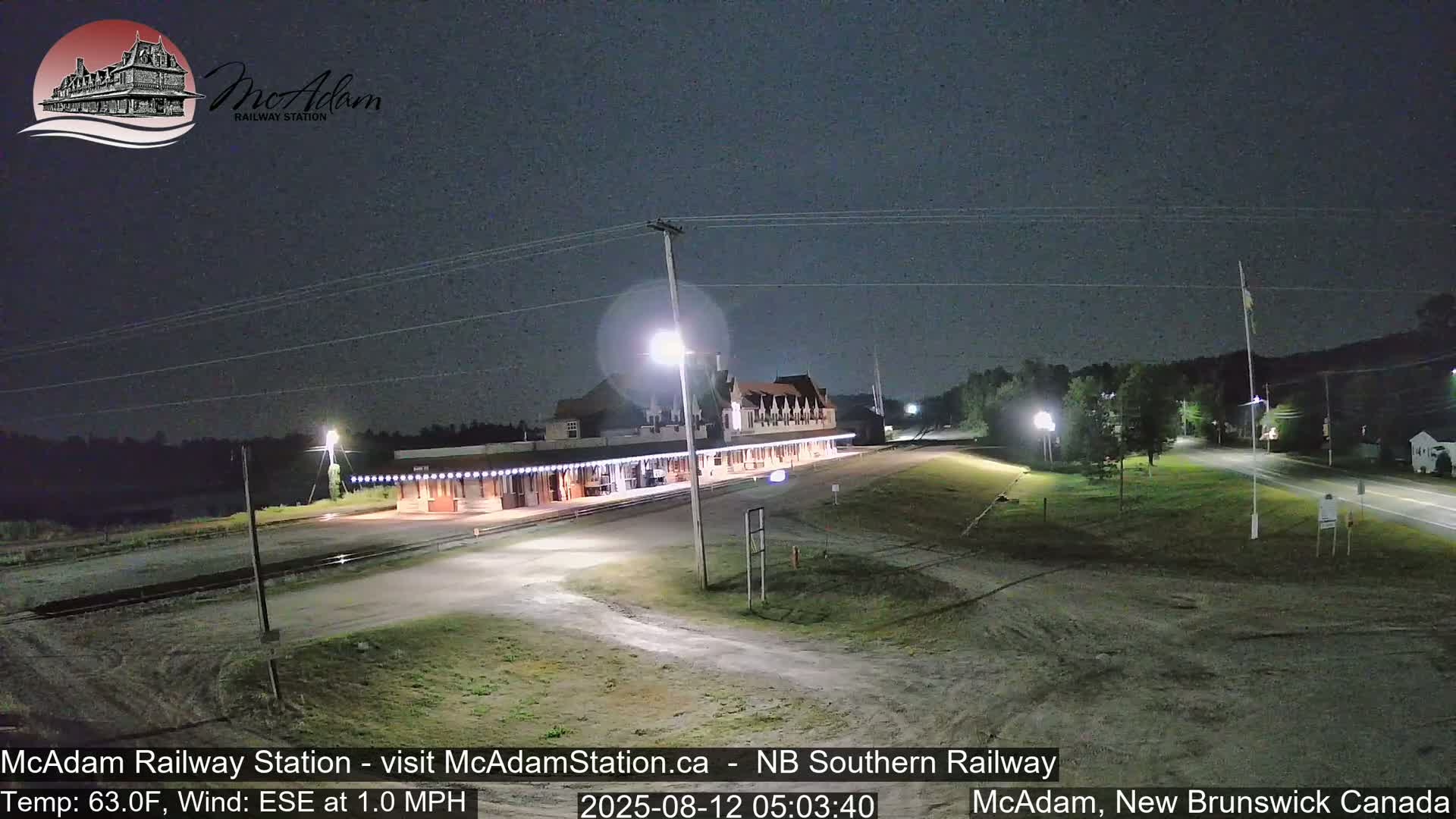 A nighttime view of a long, well-lit railway station building under a clear, dark sky with a gentle east-southeast wind.