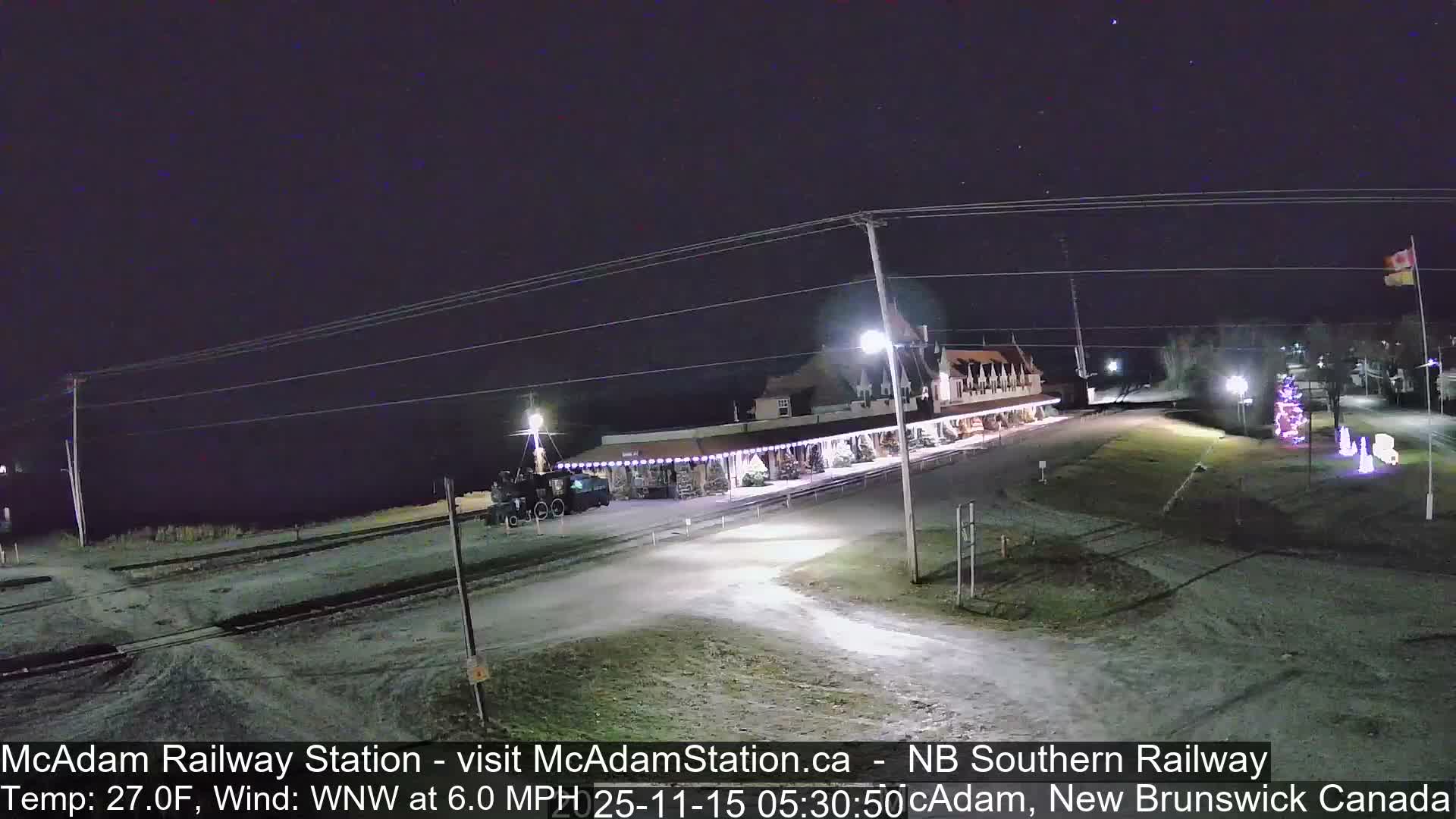 On a clear, cold night, a long railway station building adorned with festive lights sits beside tracks, with a brightly lit Christmas tree and other decorations visible on the frosted ground.
