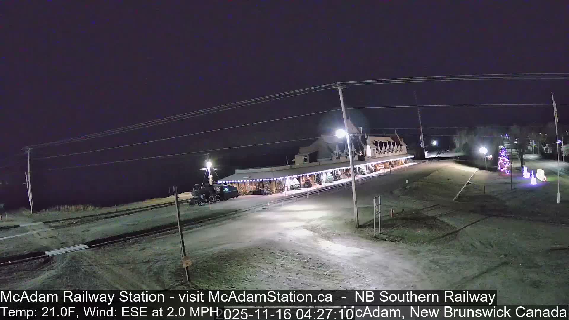 A brightly lit historic railway station, adorned with festive lights and featuring a parked locomotive on tracks, stands under a clear, cold night sky.
