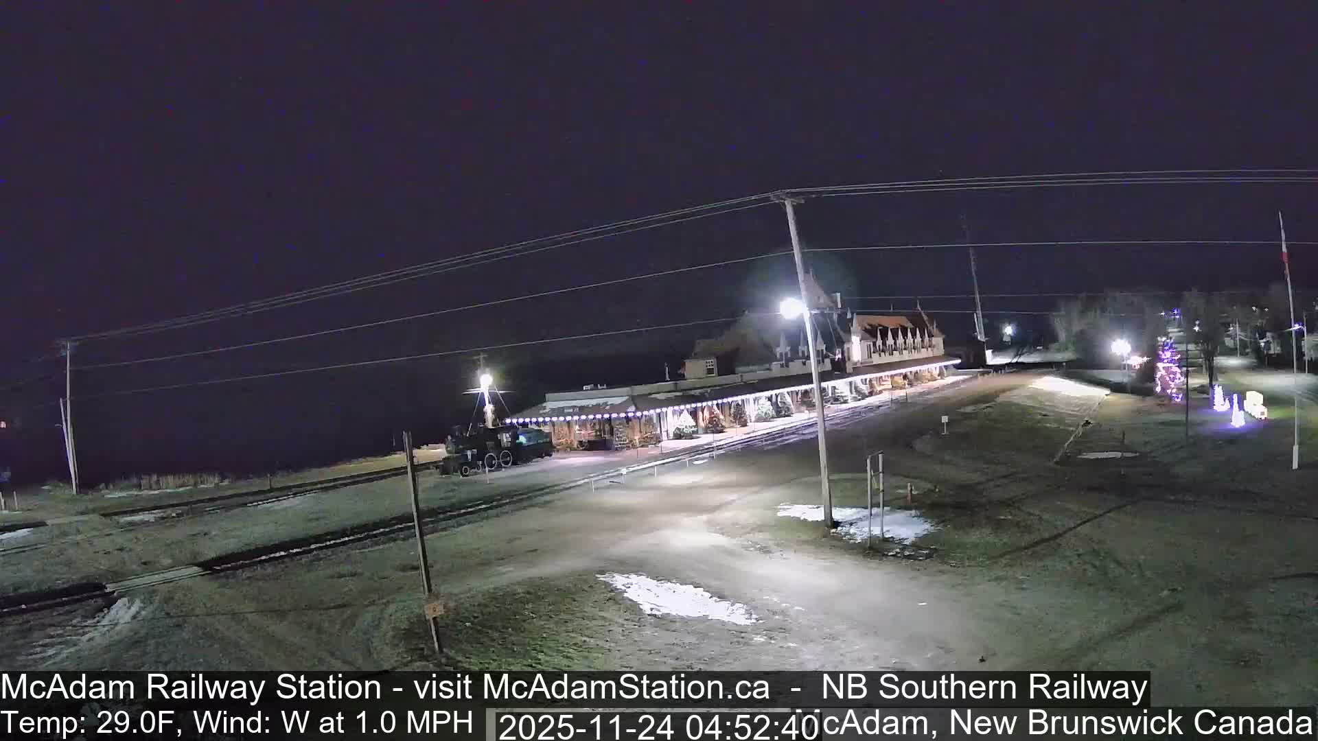 A historic railway station building, adorned with string lights, stands next to a parked train on a clear, cold night with visible patches of snow on the ground.