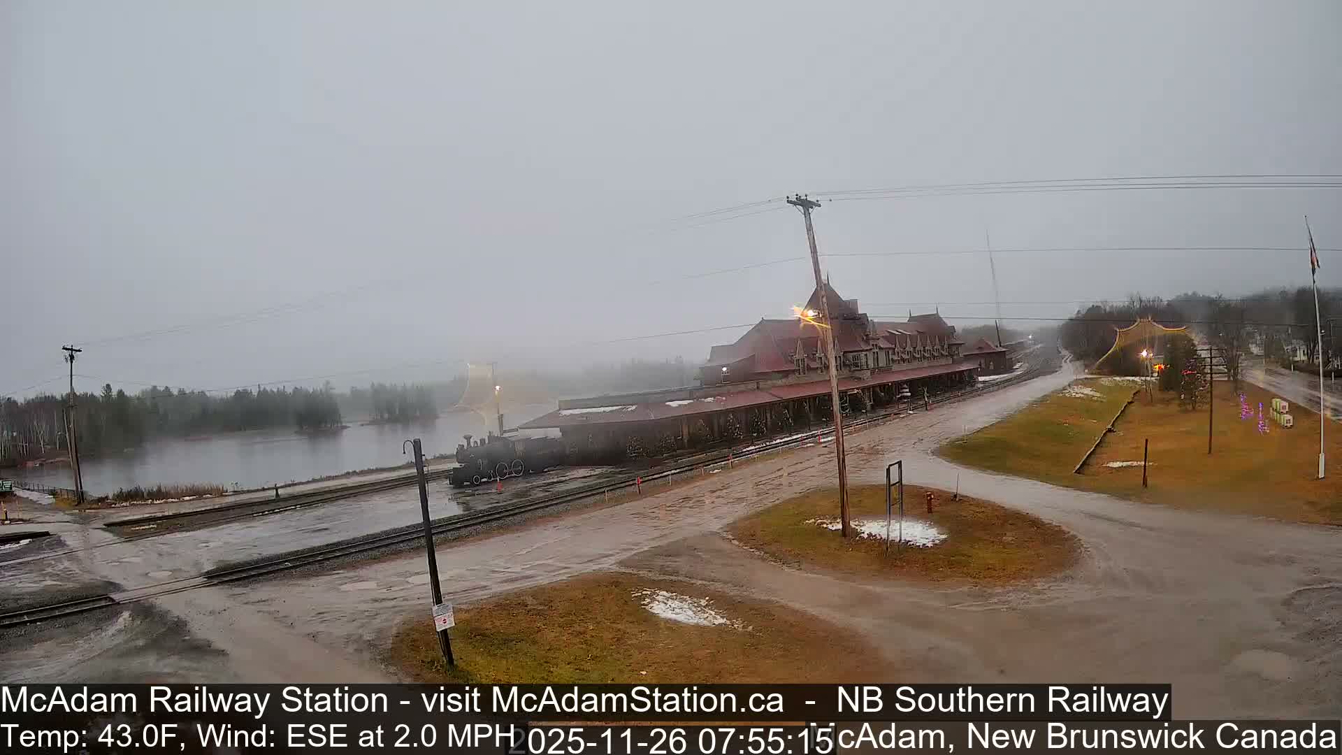 A historic railway station building, adorned with string lights, stands next to a parked train on a clear, cold night with visible patches of snow on the ground.