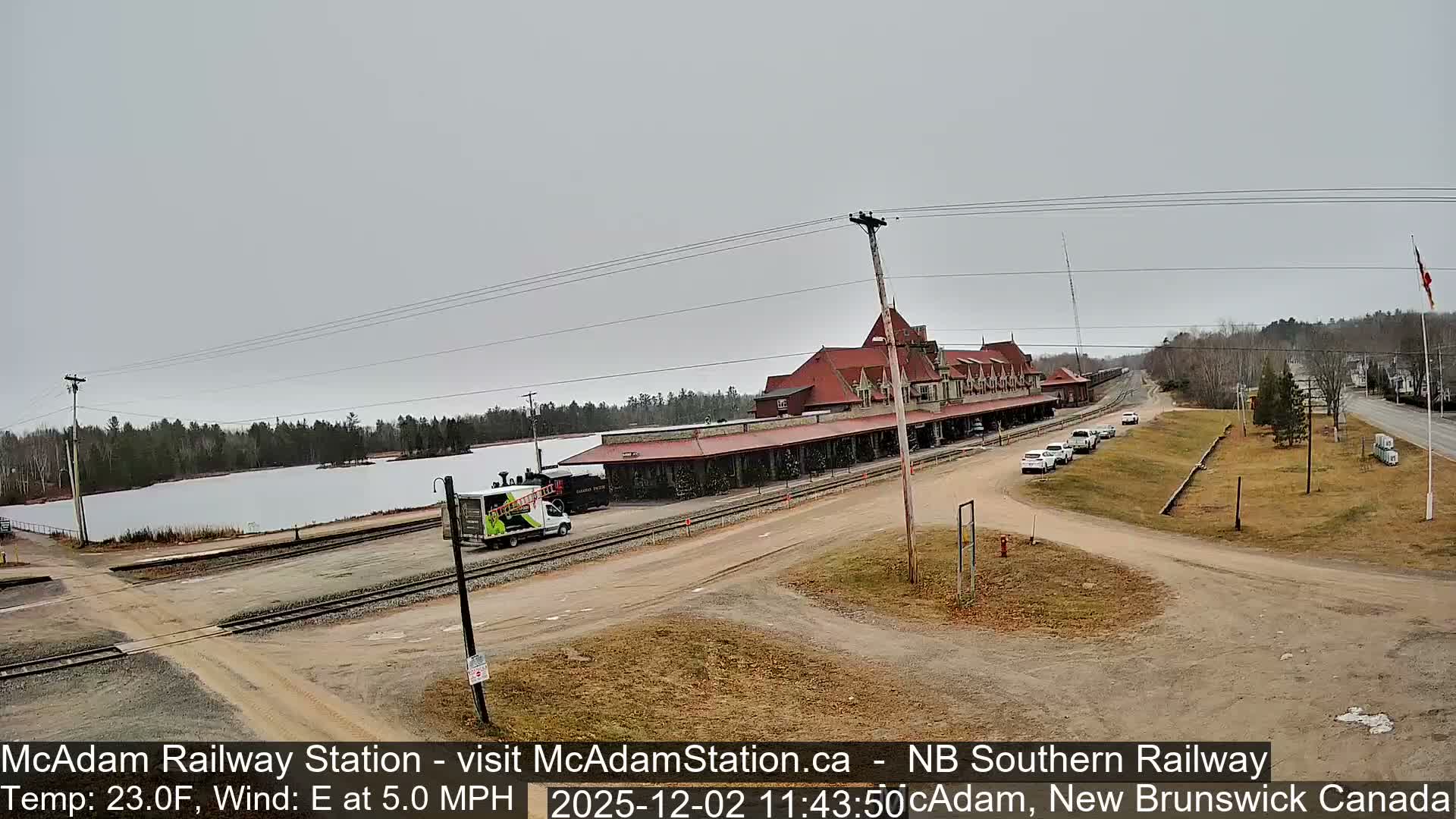 Under an overcast sky, a large, ornate red-roofed railway station stands beside a partially frozen lake and railway tracks, with a few cars and a truck visible on a cold, grey day.